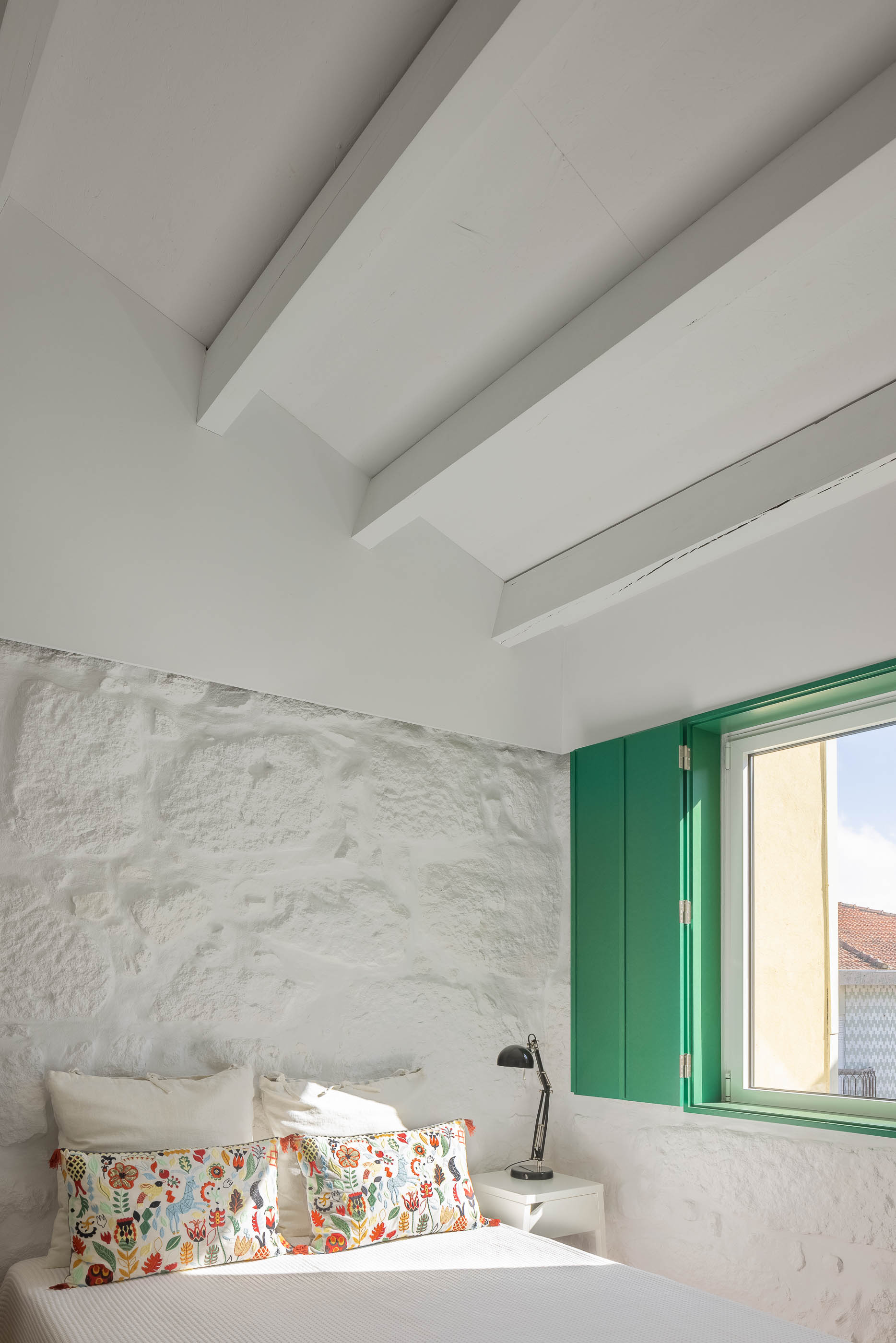 Quarto: Cama com almofadas coloridas, parede em pedra branca, vigas de madeira pintadas de branco e portadas verdes. — Casa Covelo arquitetura spaceworkers em Porto