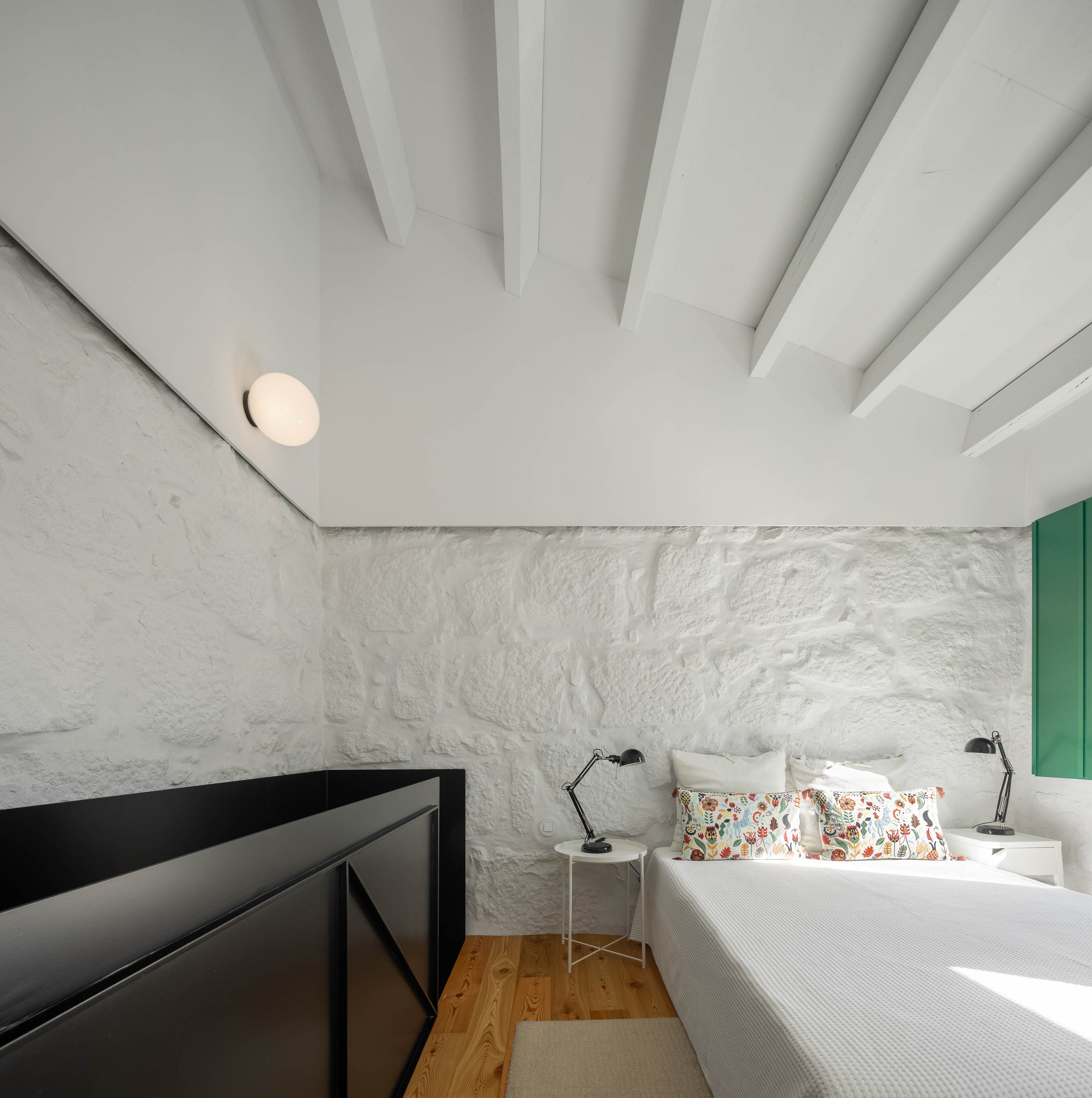 Quarto: Cama, mesas de cabeceira brancas, parede em pedra branca, vigas de madeira e guarda-corpo preto com padrão geométrico. — Casa Covelo arquitetura spaceworkers em Porto