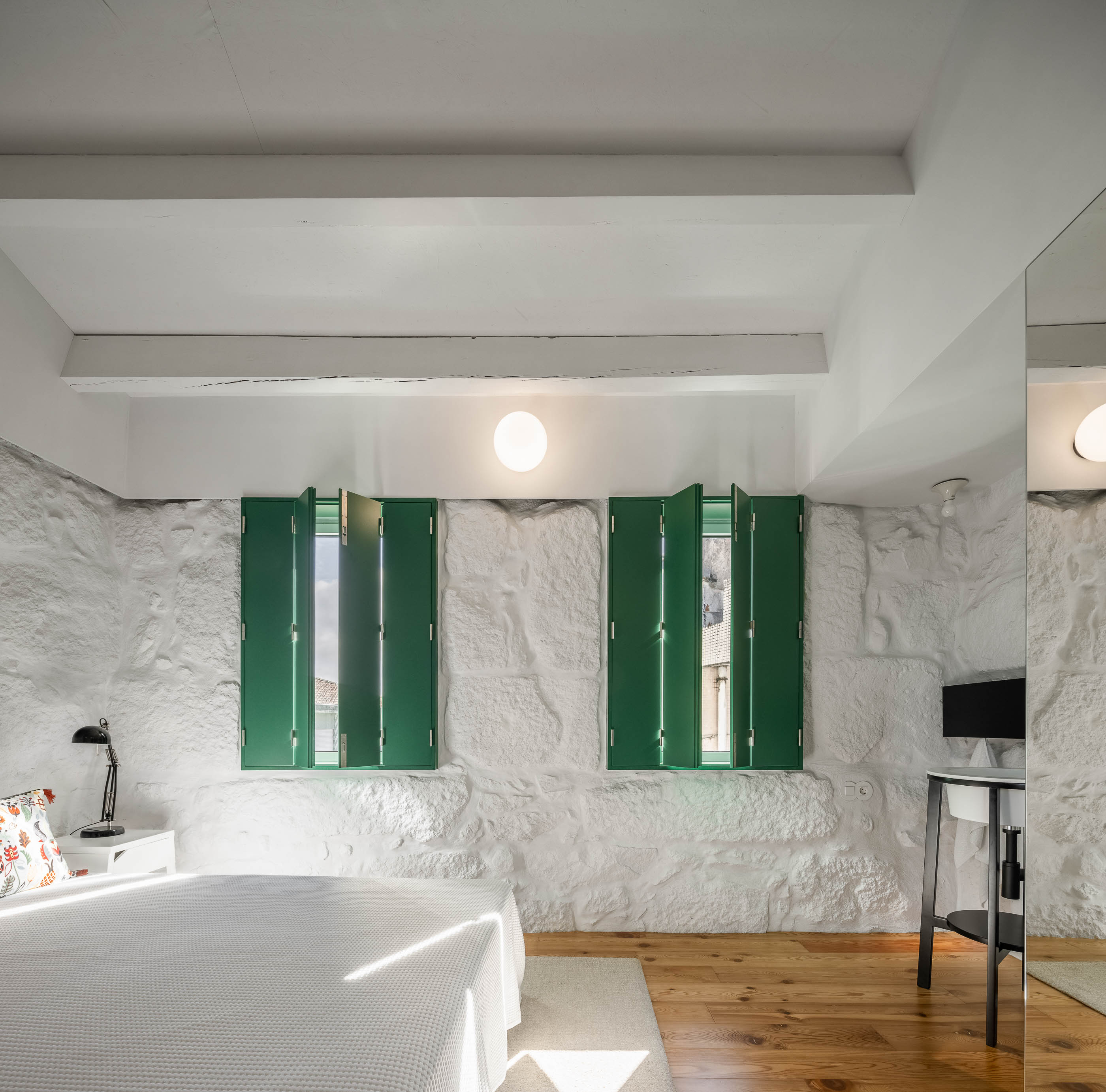 Quarto: Cama com roupa branca, parede em pedra branca, portadas verdes e espelho amplo. Pavimento em madeira clara. — Casa Covelo arquitetura spaceworkers em Porto