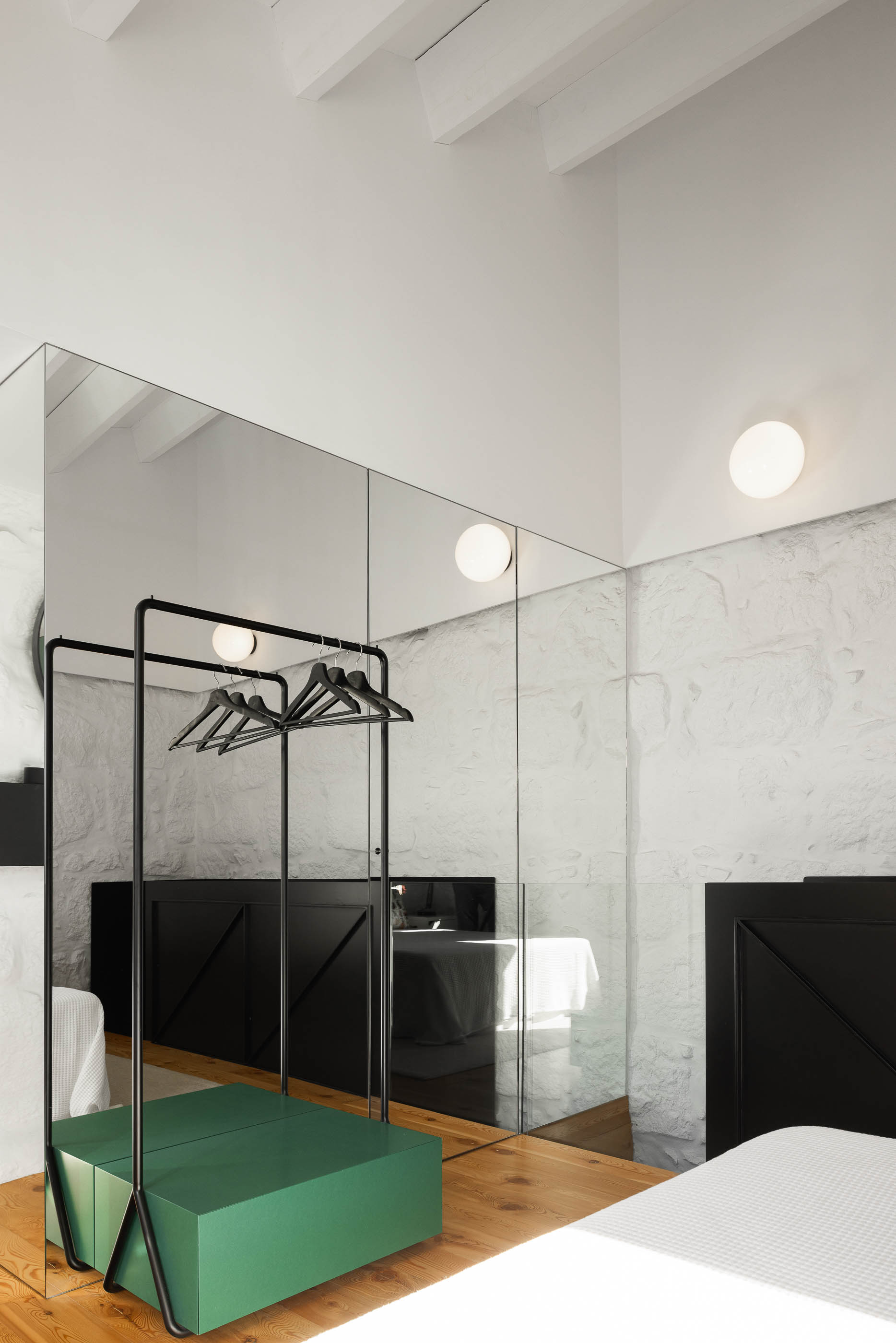 Quarto: Espelho amplo, estrutura de roupa preta, arca verde e pavimento em madeira clara. Paredes em pedra branca. — Casa Covelo arquitetura spaceworkers em Porto