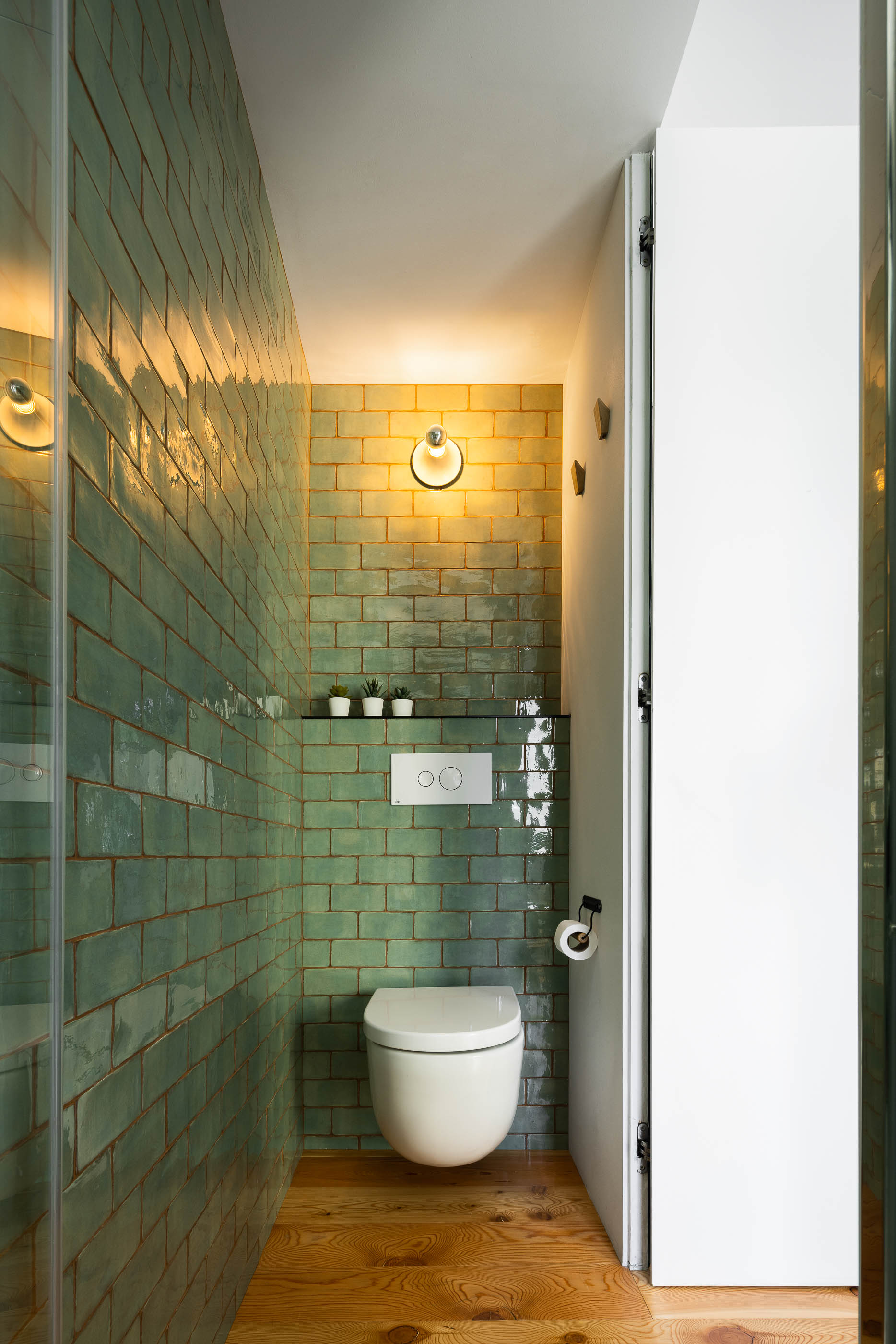 Casa de banho: Azulejos verdes brilhantes, sanita branca e pavimento em madeira clara. Candeeiro de parede. — Casa Covelo arquitetura spaceworkers em Porto
