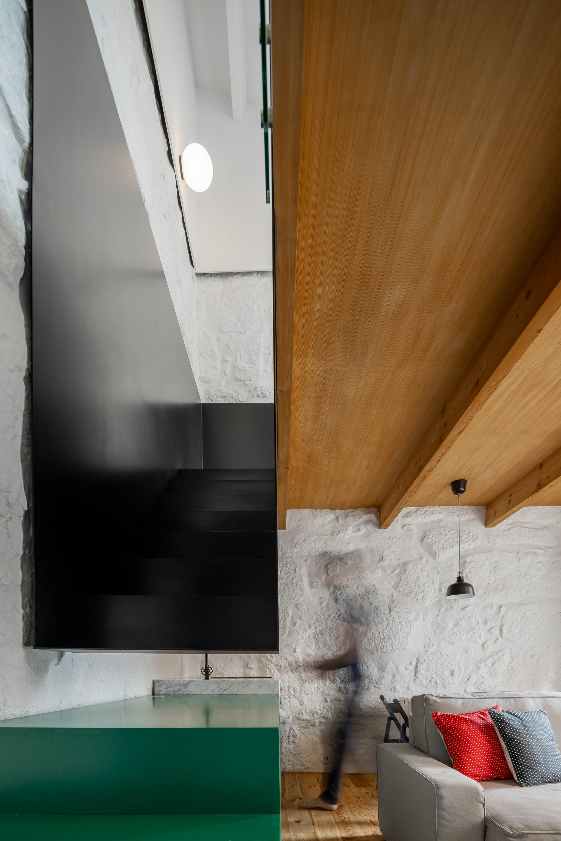 Sala de estar/Escada: Escada preta, parede em pedra branca, teto em madeira e sofá cinzento com almofadas coloridas. — Casa Covelo arquitetura spaceworkers em Porto