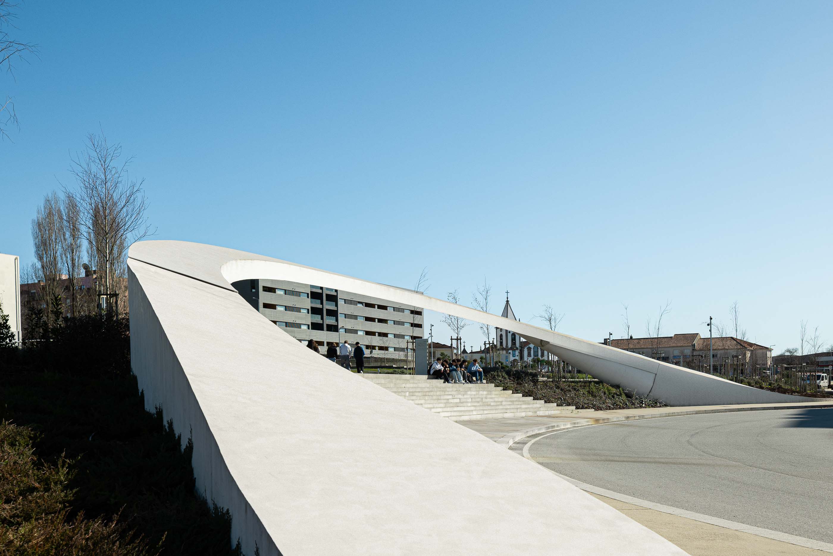 Parque: Passadiço curvo em betão que atravessa a rotunda, com escadaria e pessoas a circular. — Parque Urbano Gondomar arquitetura Paulo Merlini Architects em Gondomar