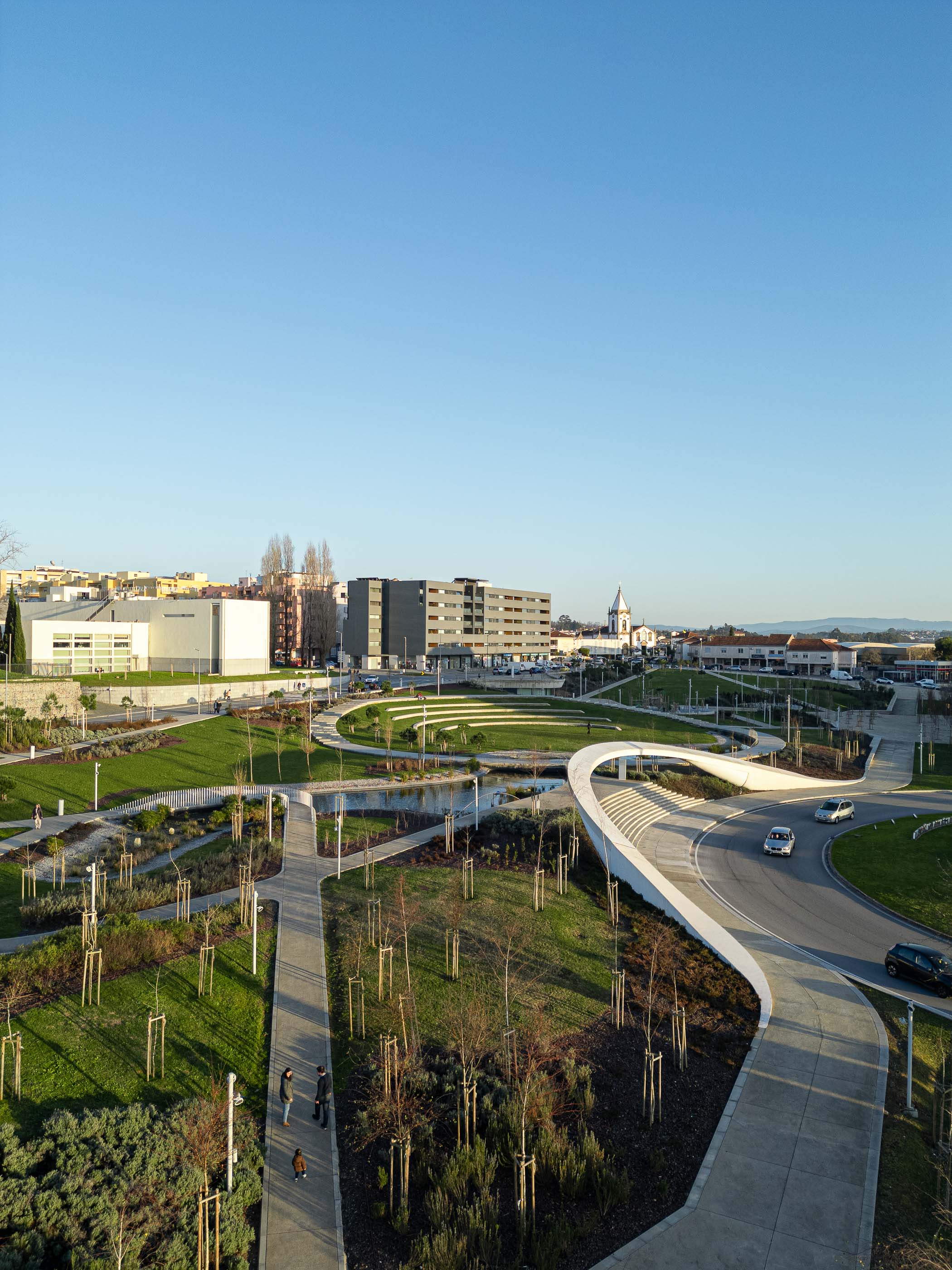 Parque: Vista geral do parque com passadiço curvo, lago artificial e edifícios na envolvente. — Parque Urbano Gondomar arquitetura Paulo Merlini Architects em Gondomar