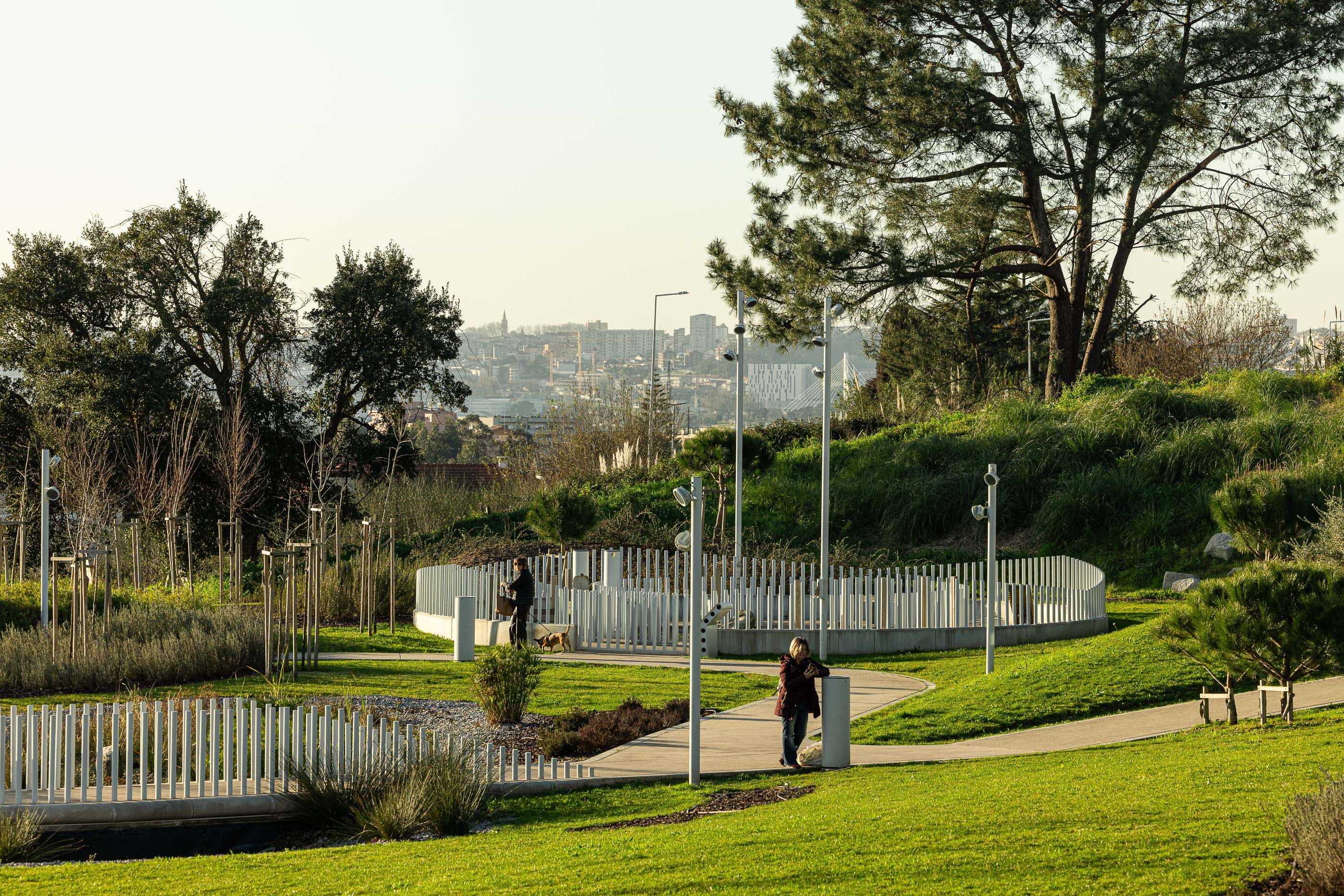 Parque: Caminho pedonal delimitado por elementos verticais em metal branco, com vegetação e árvores. — Parque Urbano Gondomar arquitetura Paulo Merlini Architects em Gondomar