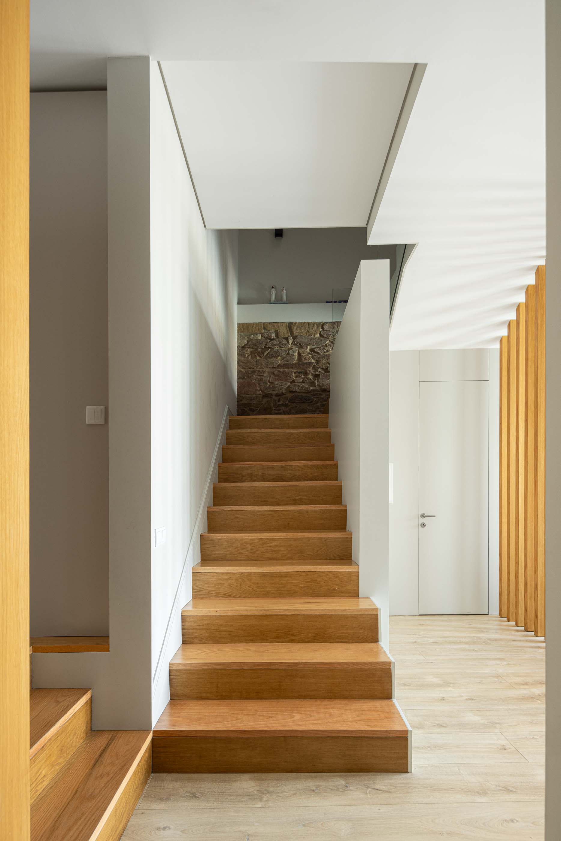 Escadas: Escadas em madeira com parede em pedra exposta e porta branca minimalista. — Casa do Parque IV arquitetura Ricardo Azevedo Arquitecto em Santo Tirso
