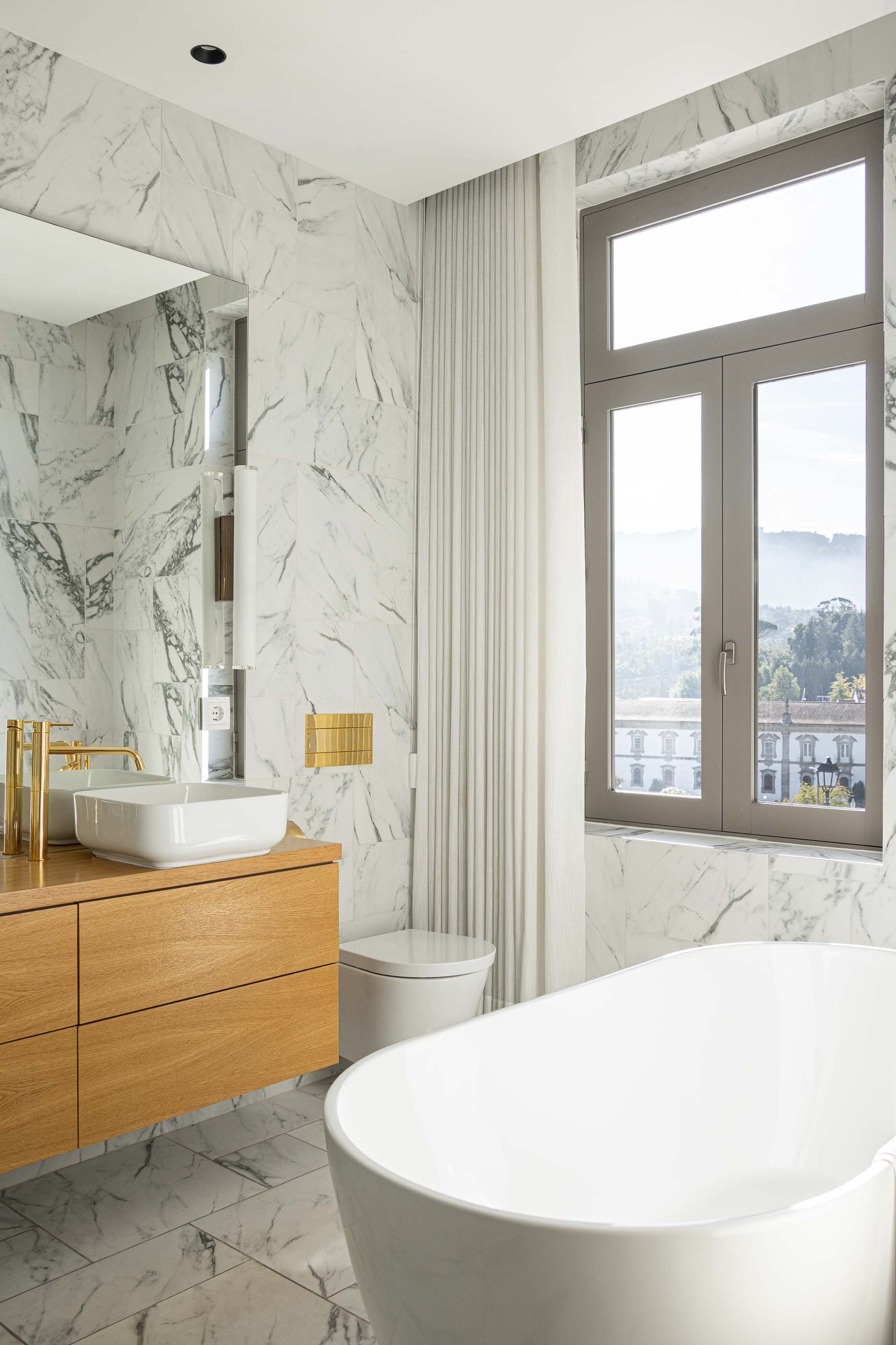 Casa de banho: Casa de banho com lavatório branco sobre móvel em madeira, banheira branca e revestimento em mármore. — Casa do Parque IV arquitetura Ricardo Azevedo Arquitecto em Santo Tirso