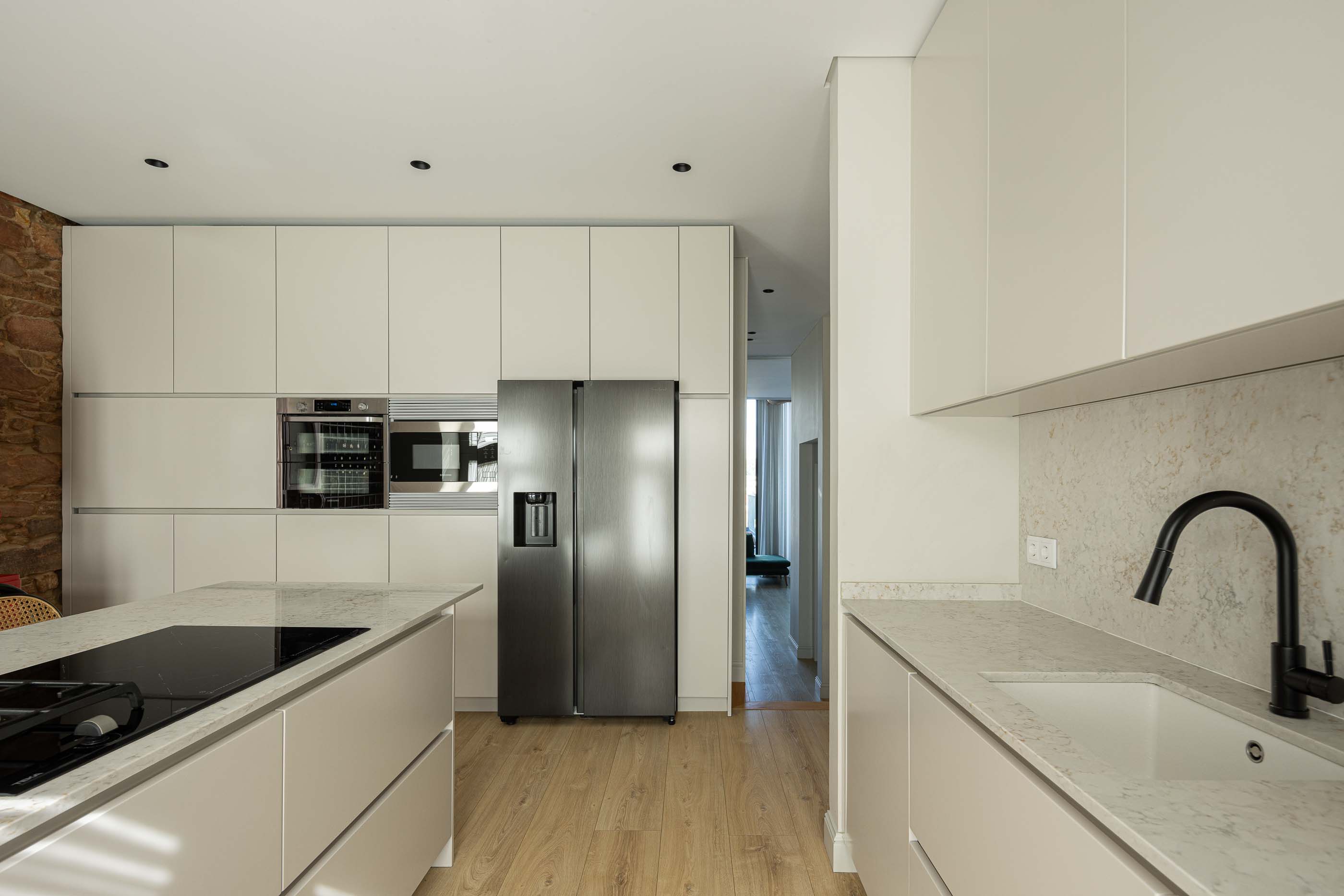 Cozinha: Cozinha com ilha, armários brancos, eletrodomésticos em inox e parede em pedra exposta. — Casa do Parque IV arquitetura Ricardo Azevedo Arquitecto em Santo Tirso