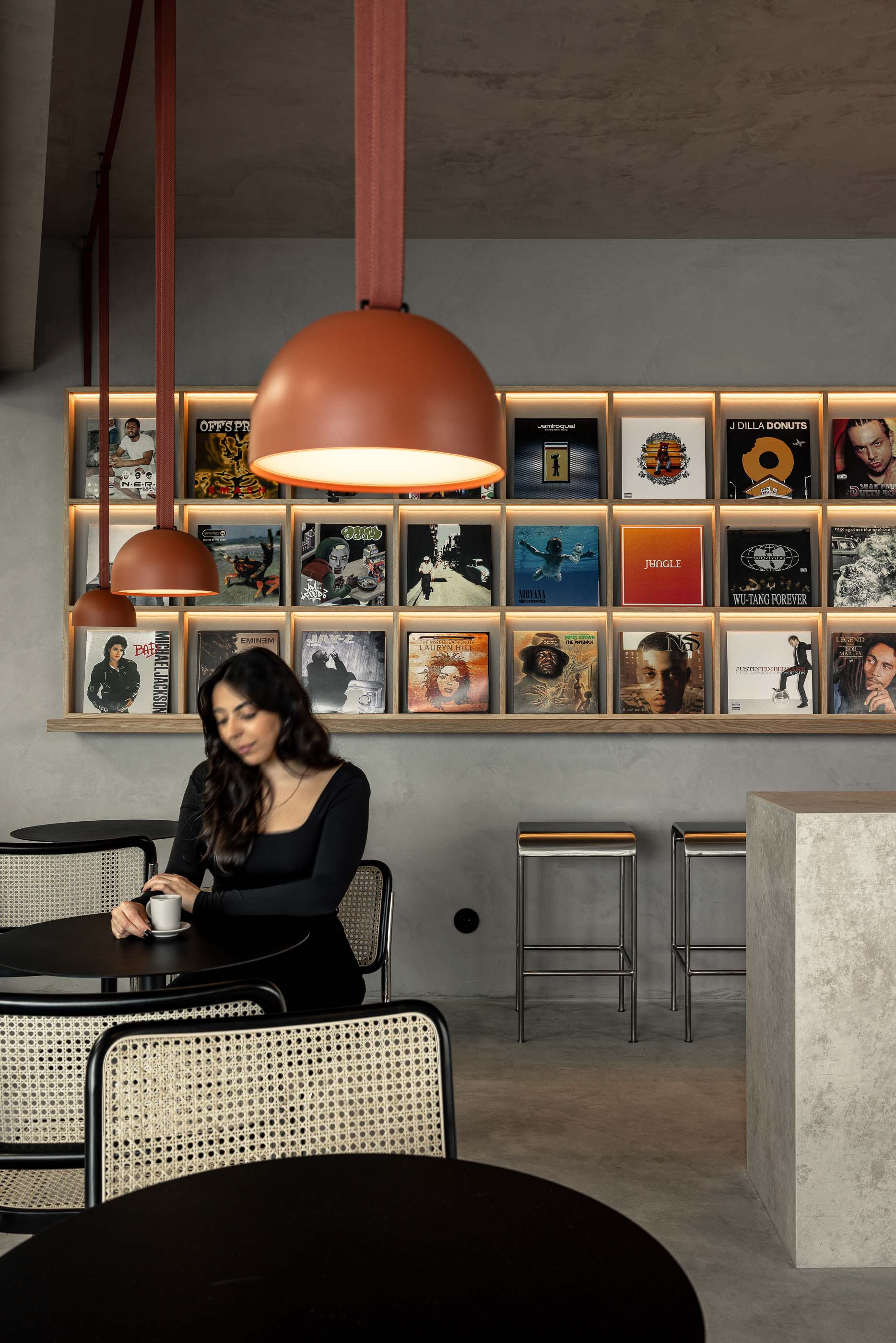 Zona de mesas: Candeeiros cor de tijolo sobre mesas redondas com cadeiras de palhinha, pavimento cinzento e parede texturada. — KI.art.music.coffee arquitetura MAT Arquitetura e Interiores em Porto