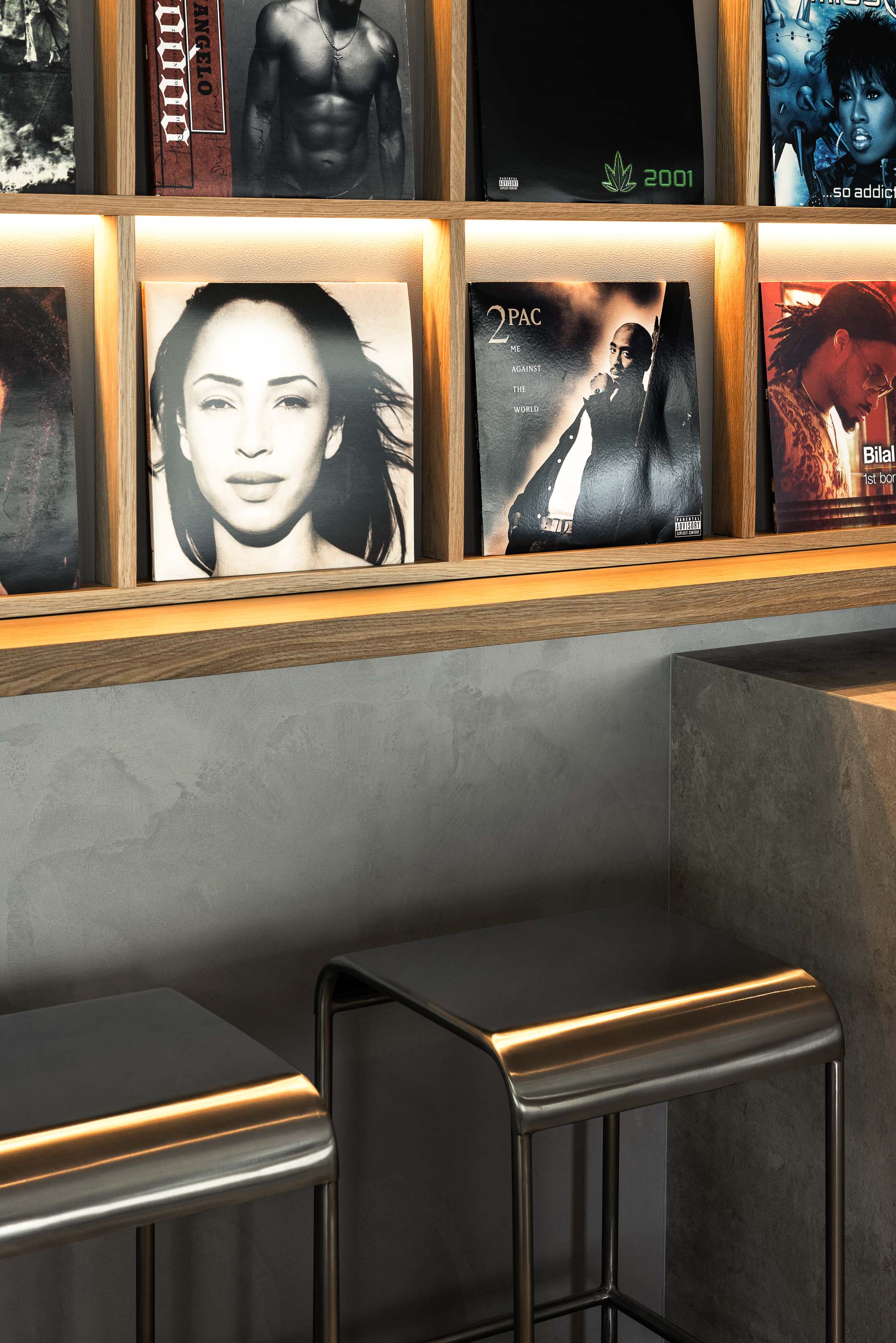 Detalhe da estante: Prateleiras de madeira iluminadas com capas de discos de vinil, sobre bancada de betão cinzento e cadeiras metálicas. — KI.art.music.coffee arquitetura MAT Arquitetura e Interiores em Porto