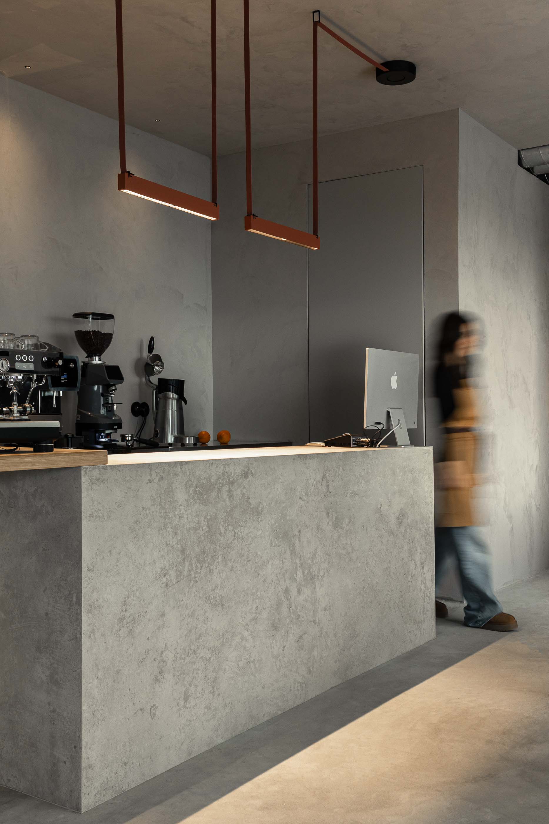 Balcão: Balcão de betão cinzento com máquina de café e computador, sob candeeiros suspensos e parede texturada. — KI.art.music.coffee arquitetura MAT Arquitetura e Interiores em Porto