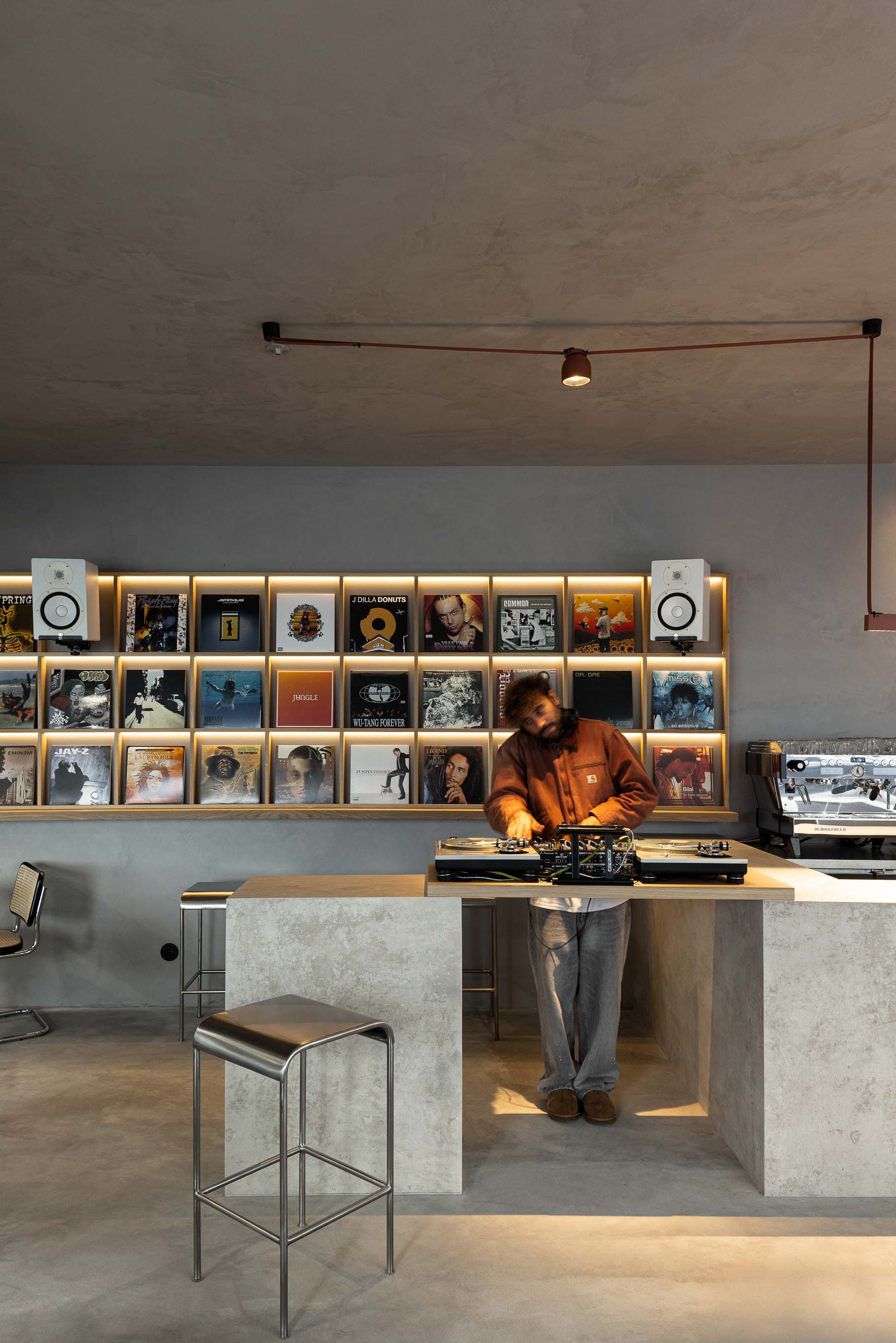 Perspectiva geral: Balcão de betão, estante de discos iluminada, cadeiras metálicas e pavimento cinzento. — KI.art.music.coffee arquitetura MAT Arquitetura e Interiores em Porto