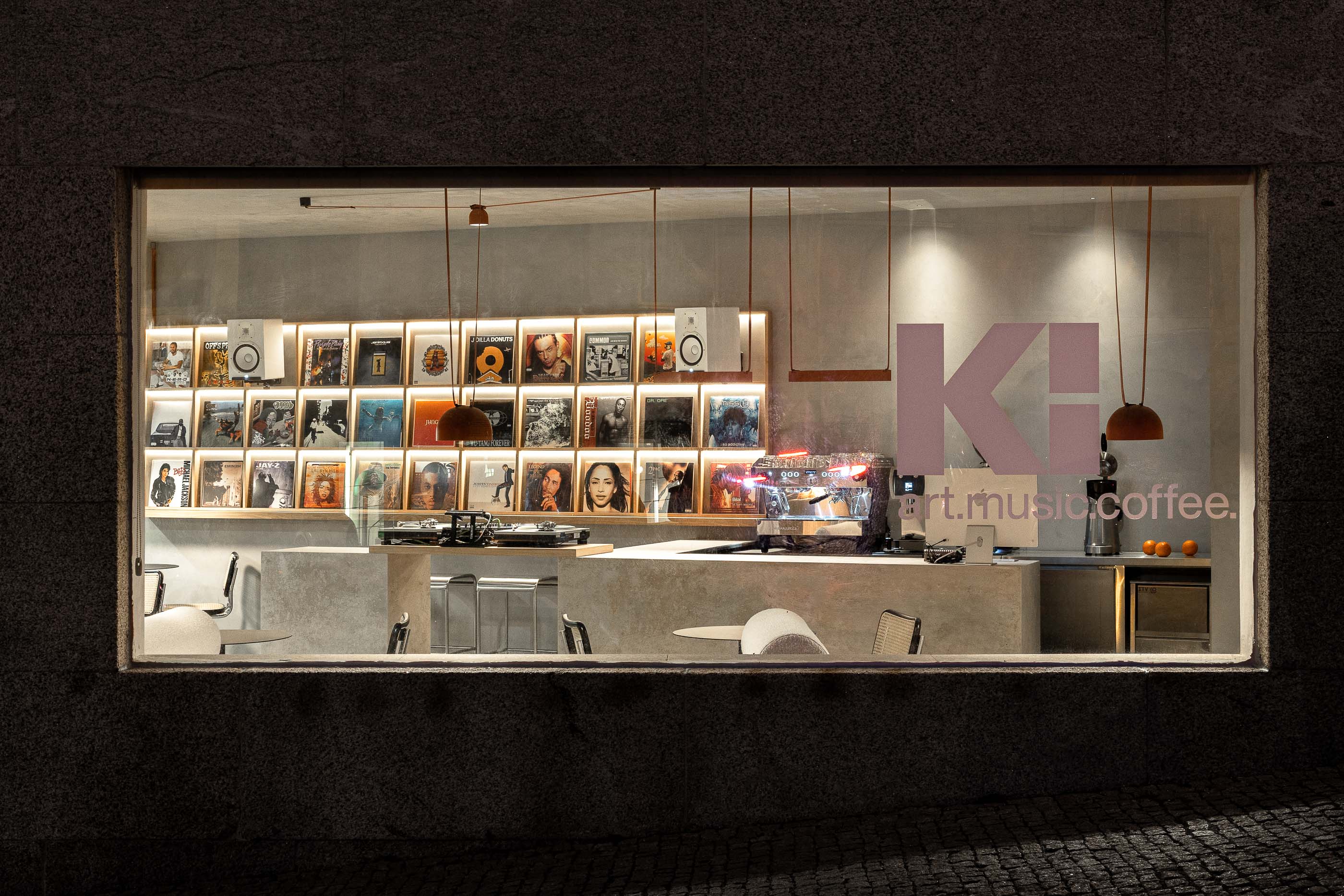 Fachada: Espaço interior visível através da montra, com estante de discos, balcão de betão e sofás. — KI.art.music.coffee arquitetura MAT Arquitetura e Interiores em Porto