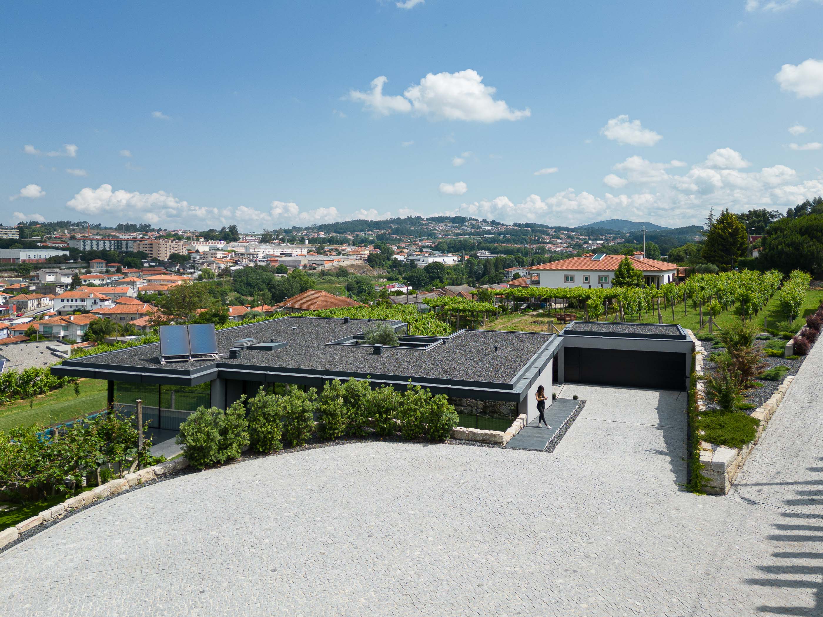 Exterior: Vista da casa com garagem, acesso em calçada, vinha e paisagem urbana ao fundo. Arquitetura moderna com revestimento escuro. — Casa da Encosta arquitetura Ricardo Azevedo Arquitecto em Santo Tirso