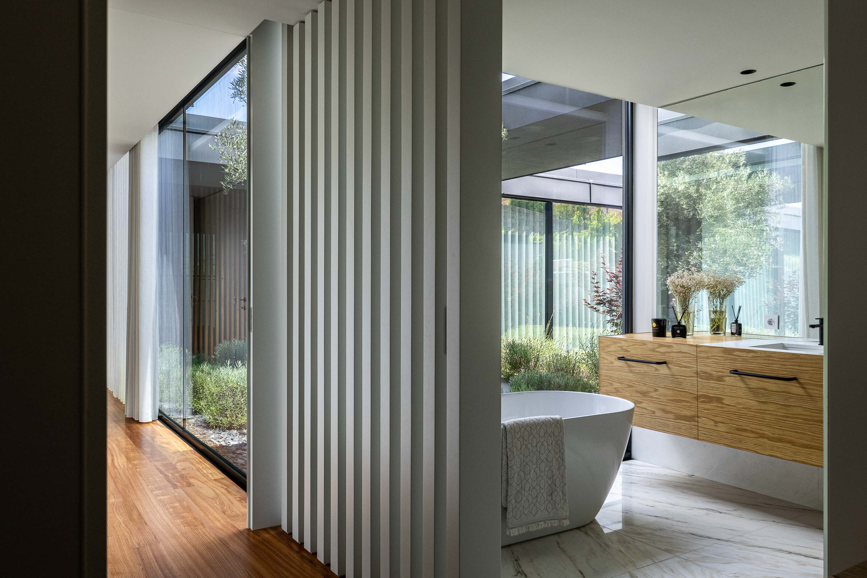 Casa de banho: Casa de banho com banheira branca, lavatório em madeira clara, divisória em ripas brancas e vista para o pátio interior. — Casa da Encosta arquitetura Ricardo Azevedo Arquitecto em Santo Tirso