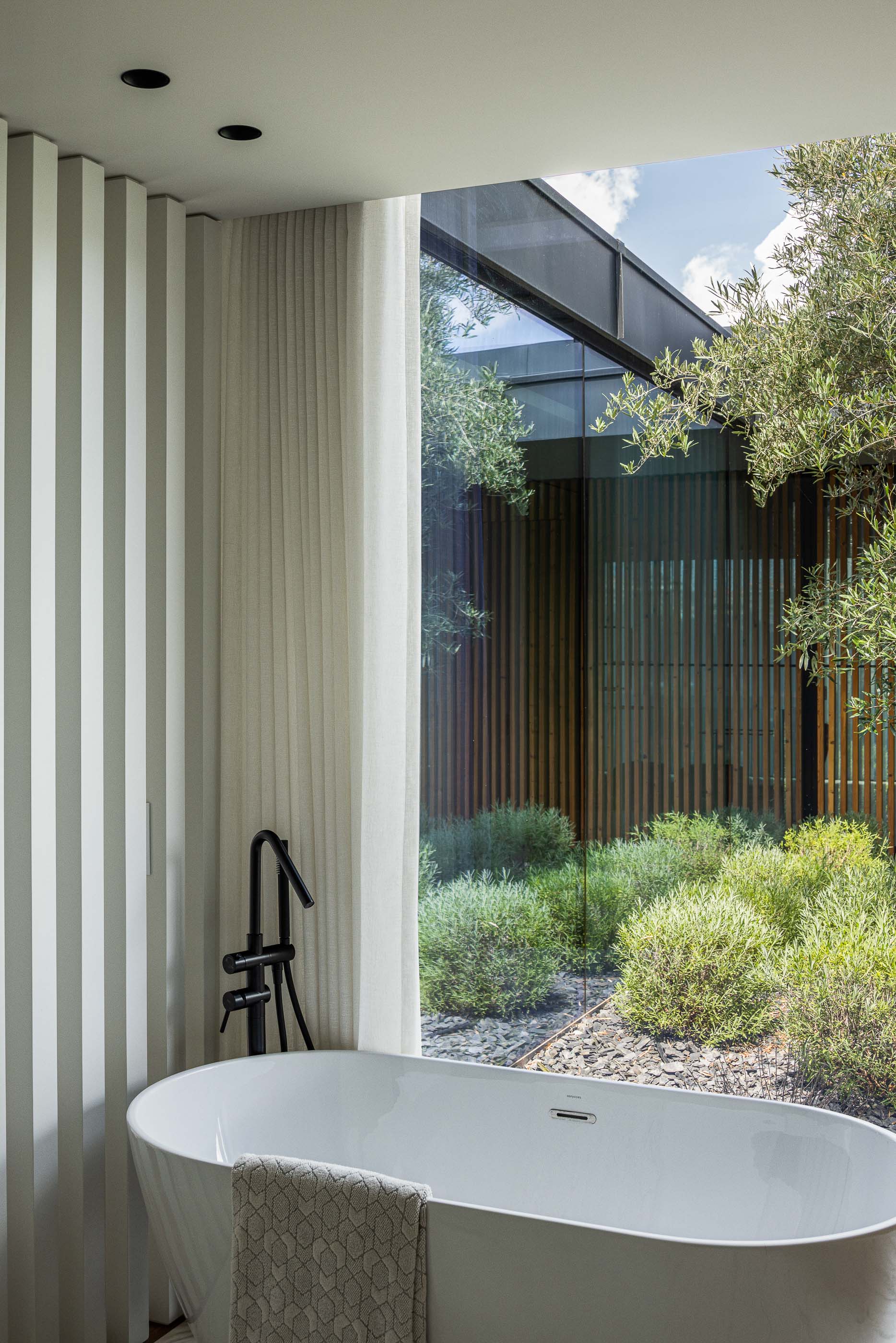 Casa de banho: Detalhe da banheira branca com torneira preta e vista para o pátio interior com oliveira. Design minimalista e elegante. — Casa da Encosta arquitetura Ricardo Azevedo Arquitecto em Santo Tirso