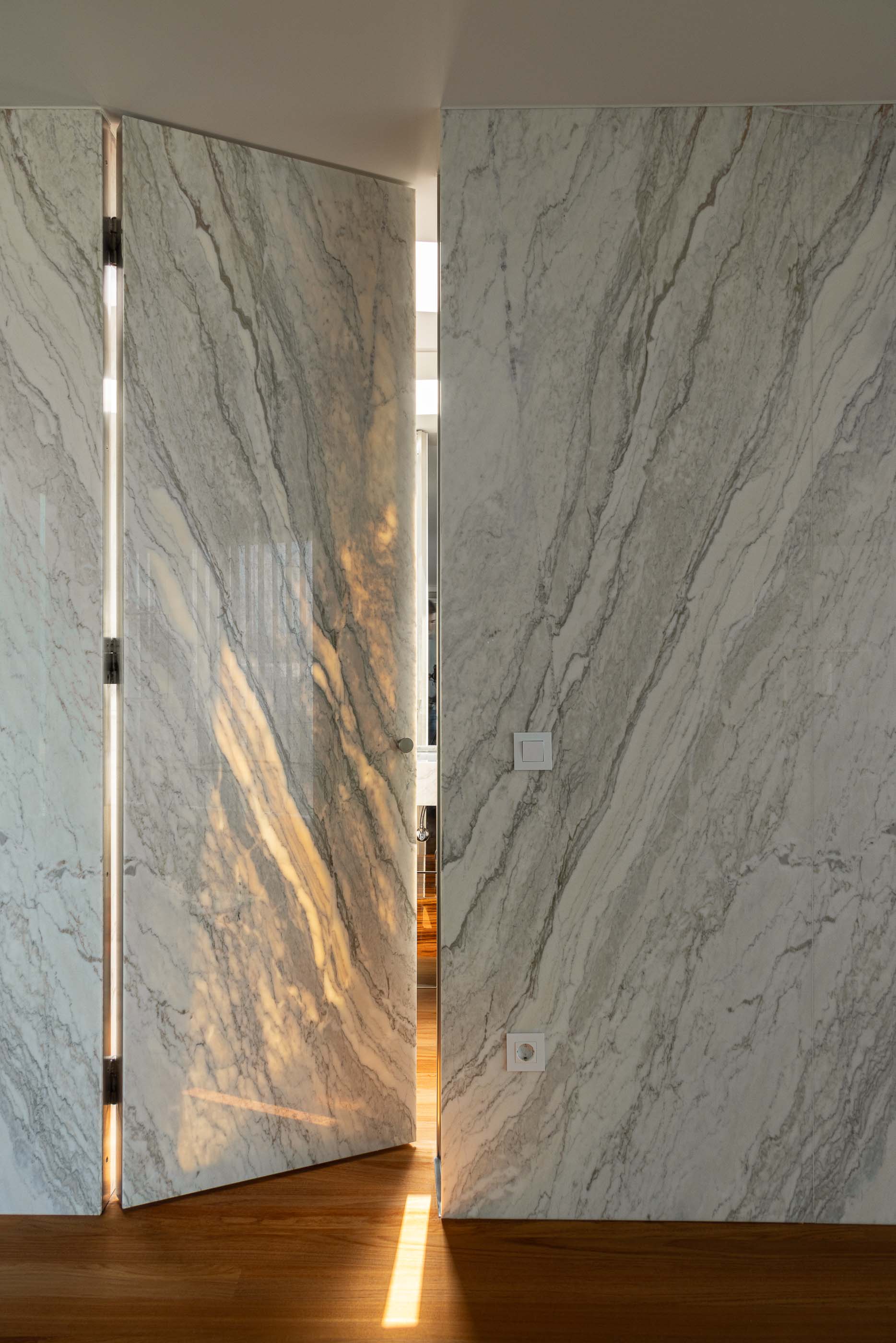 Hall de entrada: Detalhe da divisória em mármore com porta pivotante aberta, pavimento em madeira e luz natural. — Casa da Encosta arquitetura Ricardo Azevedo Arquitecto em Santo Tirso