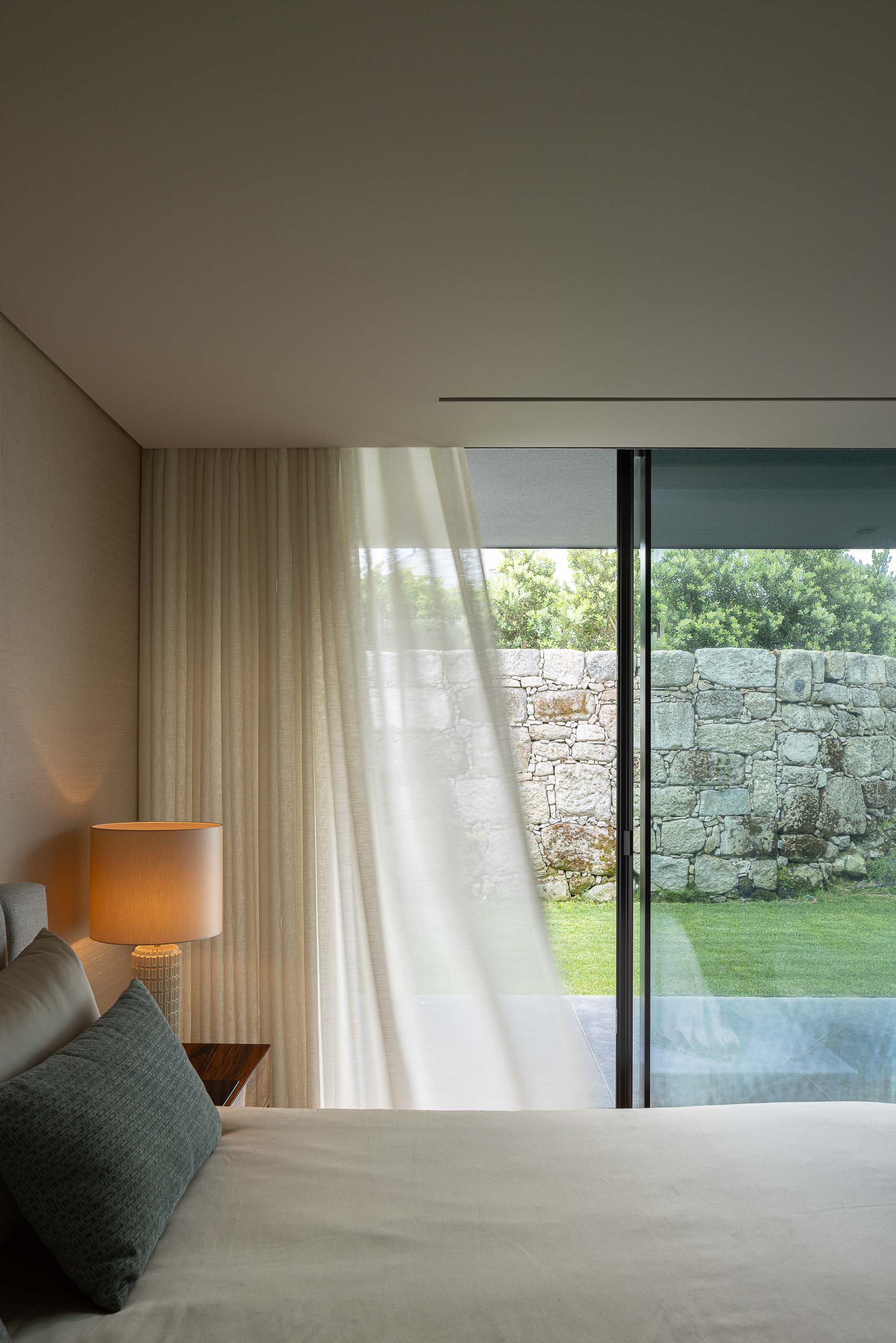 Quarto: Quarto com cama, cortinas claras e vista para o jardim através de uma janela deslizante. Decoração minimalista e relaxante. — Casa da Encosta arquitetura Ricardo Azevedo Arquitecto em Santo Tirso