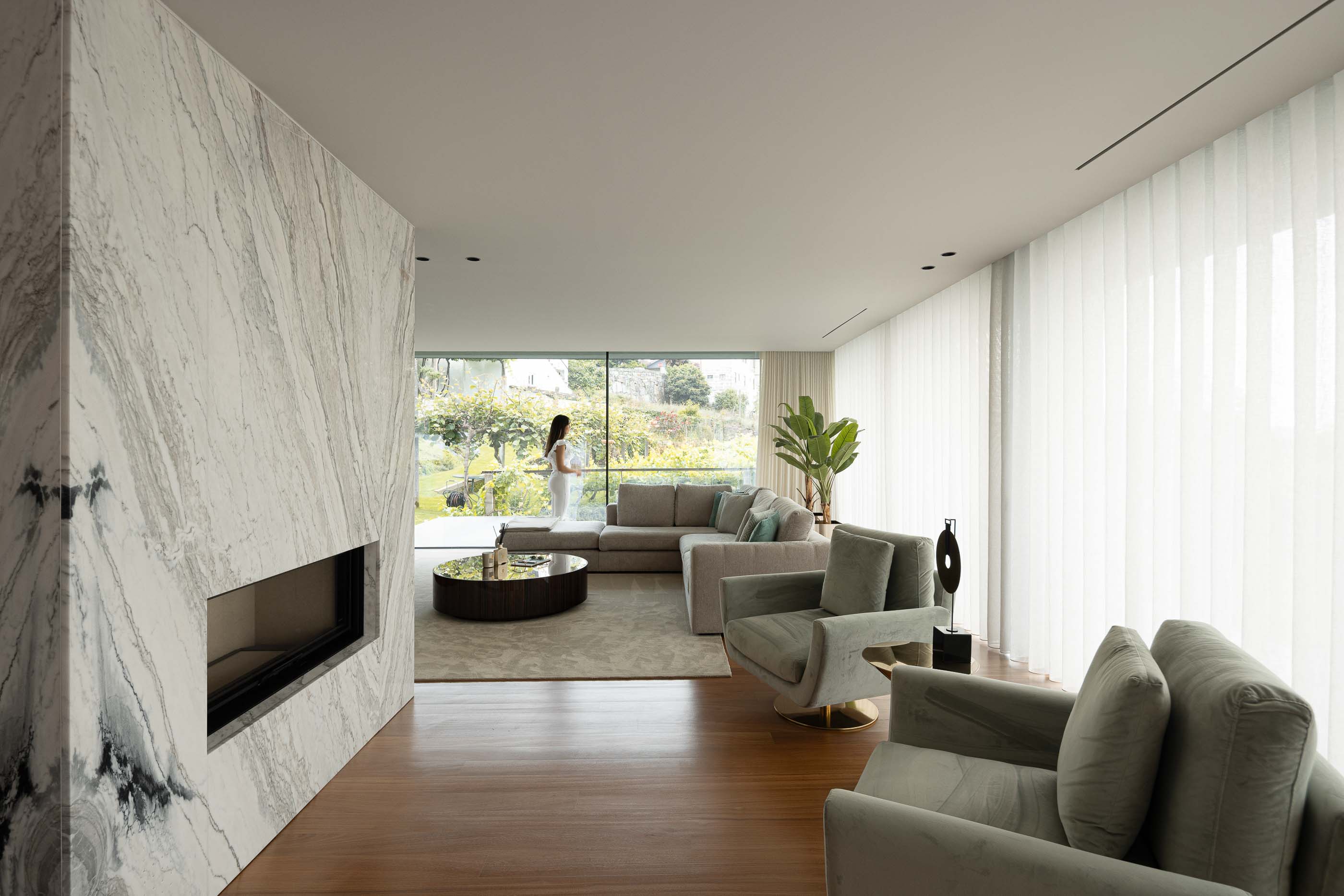 Sala de estar: Sala de estar com lareira em mármore, sofás cinzentos, alcatifa e vista para o jardim através de grandes janelas. — Casa da Encosta arquitetura Ricardo Azevedo Arquitecto em Santo Tirso