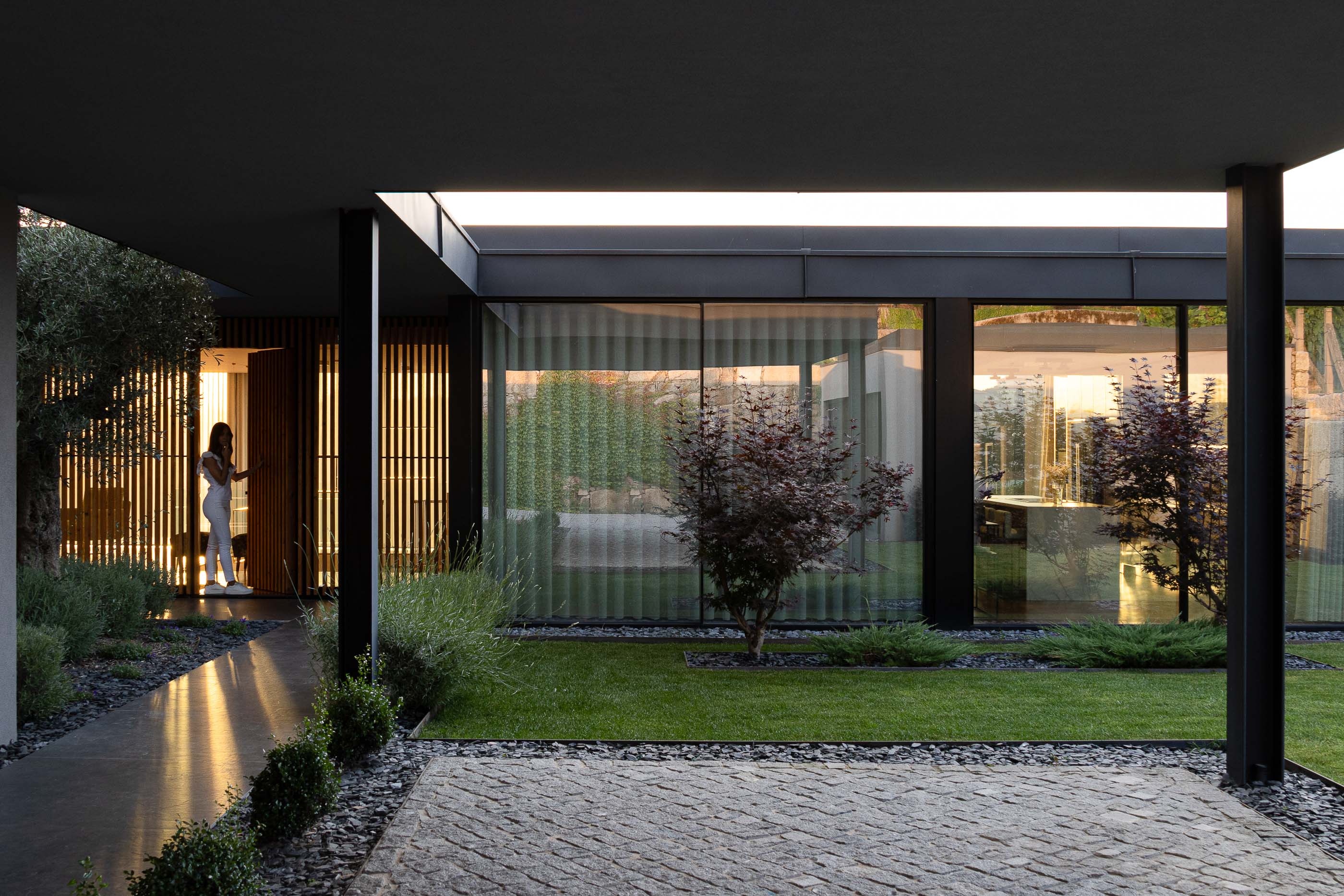 Exterior: Vista noturna da casa a partir do jardim, com iluminação indireta e pátio interior visível. Arquitetura moderna e elegante. — Casa da Encosta arquitetura Ricardo Azevedo Arquitecto em Santo Tirso