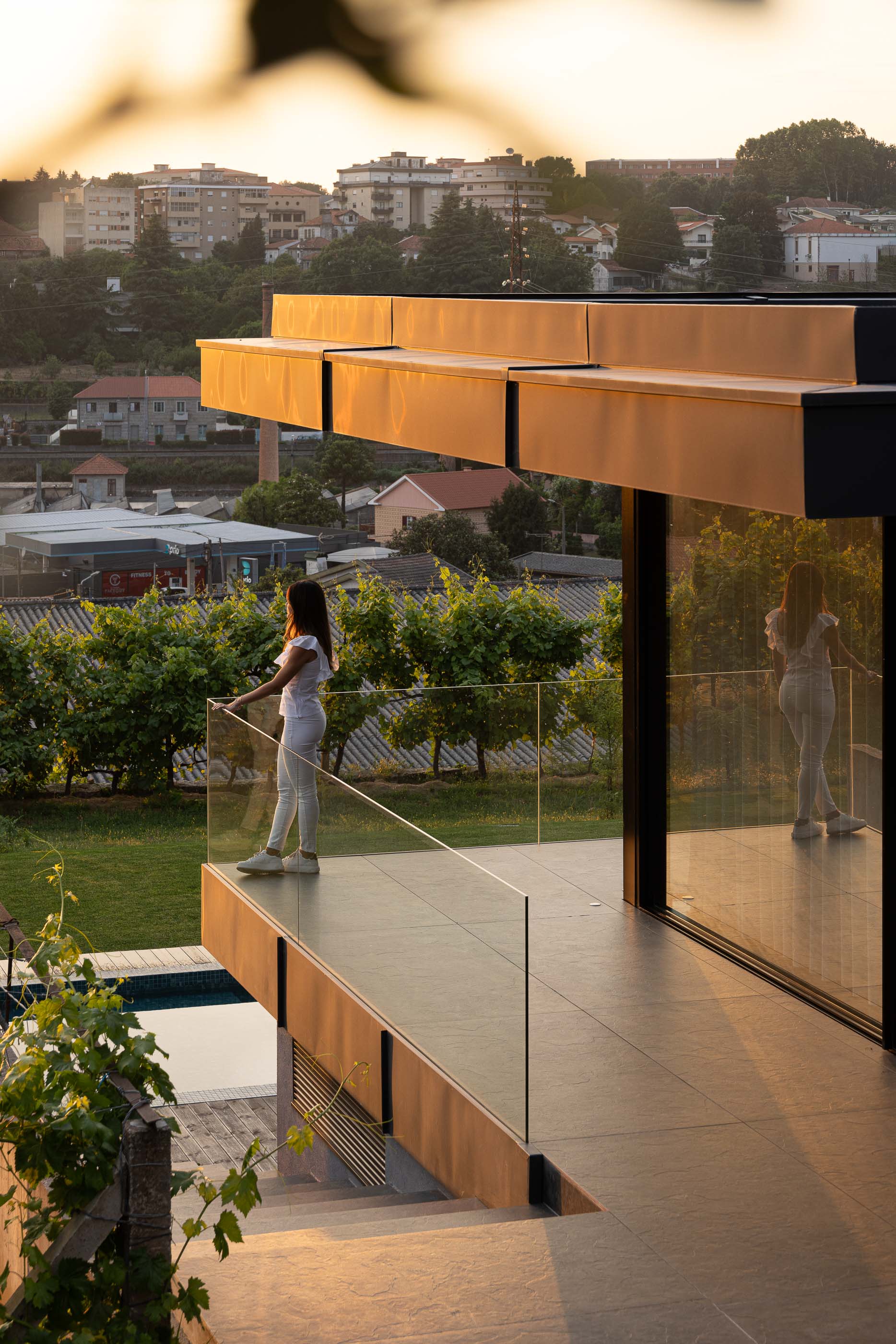 Varanda: Varanda com pavimento em cerâmica, guarda-corpo em vidro e vista para a paisagem urbana e vinha ao pôr do sol. — Casa da Encosta arquitetura Ricardo Azevedo Arquitecto em Santo Tirso