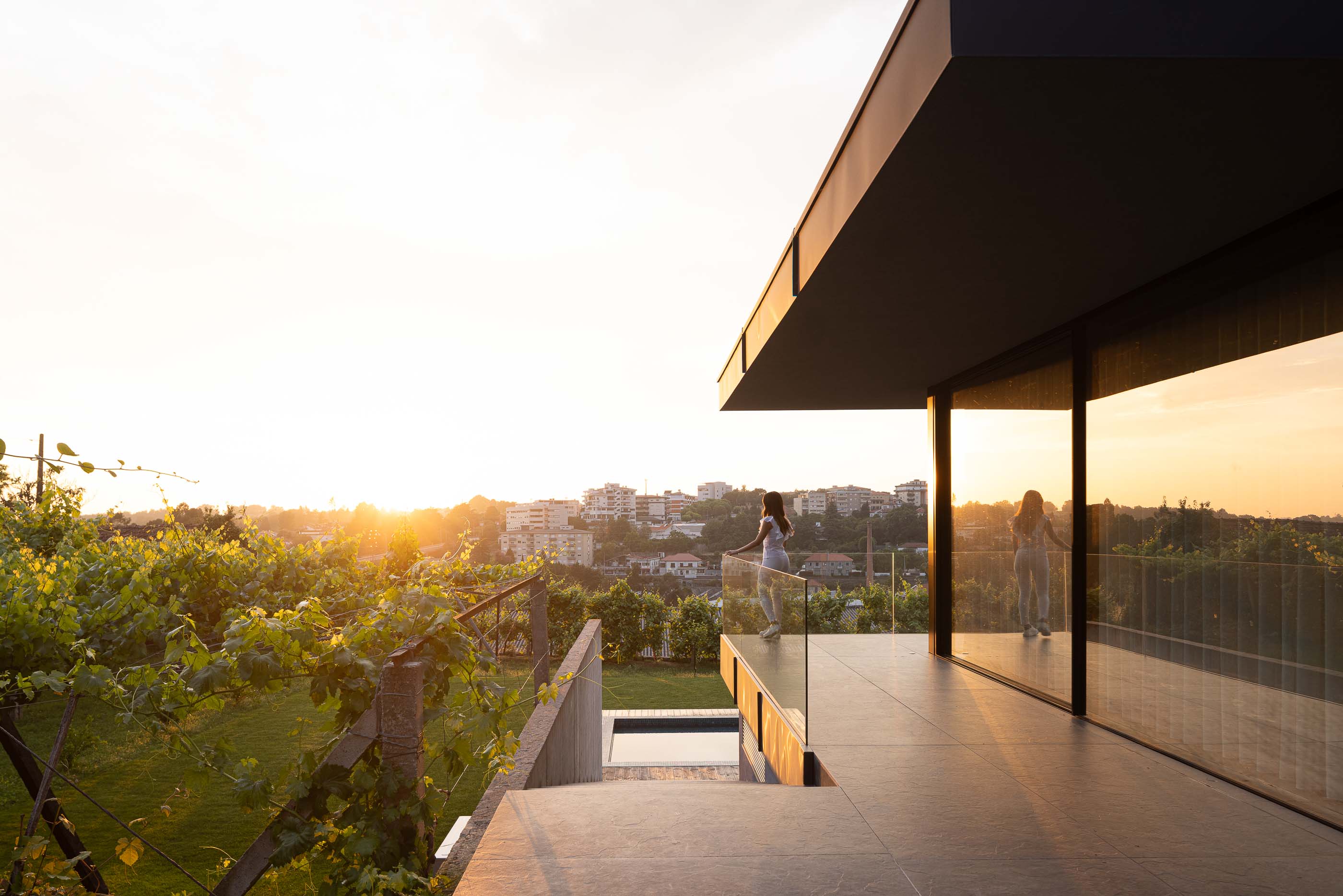 Varanda: Varanda com pavimento em cerâmica, guarda-corpo em vidro e vista para a vinha e paisagem urbana ao pôr do sol. — Casa da Encosta arquitetura Ricardo Azevedo Arquitecto em Santo Tirso