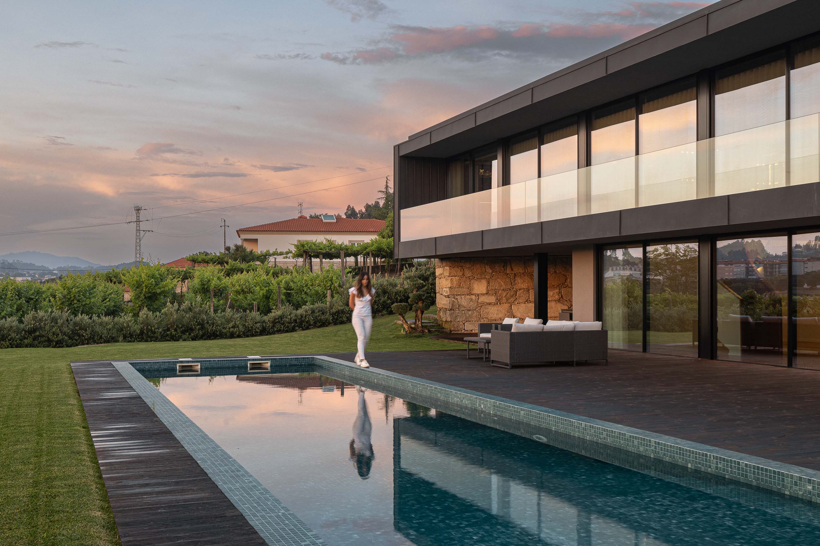 Exterior: Vista da piscina, jardim e fachada da casa com revestimento em pedra e zona de estar exterior. Arquitetura moderna integrada na paisagem. — Casa da Encosta arquitetura Ricardo Azevedo Arquitecto em Santo Tirso