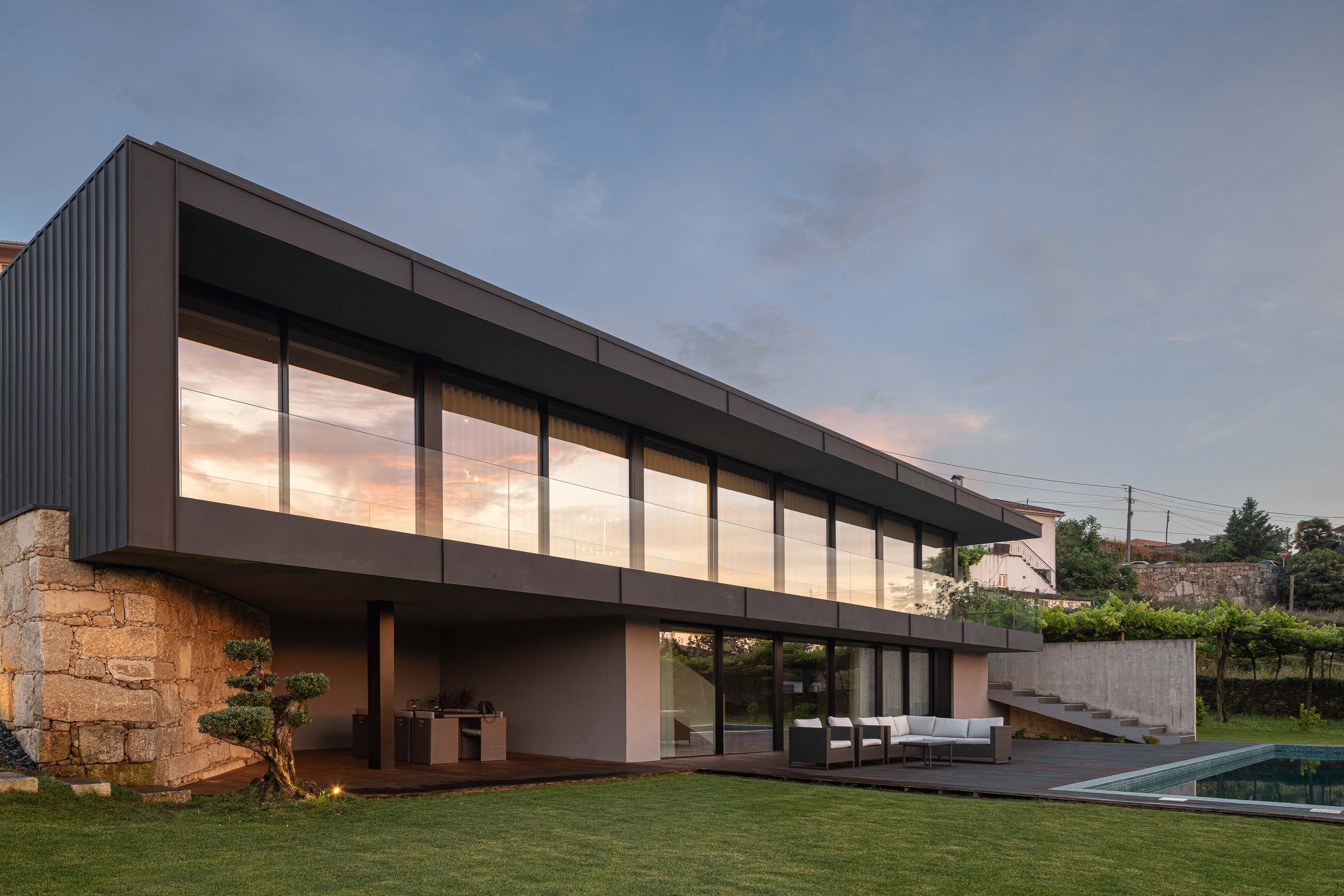 Exterior: Vista da fachada da casa com revestimento em pedra, zona de estar exterior e piscina. Arquitetura moderna e minimalista. — Casa da Encosta arquitetura Ricardo Azevedo Arquitecto em Santo Tirso