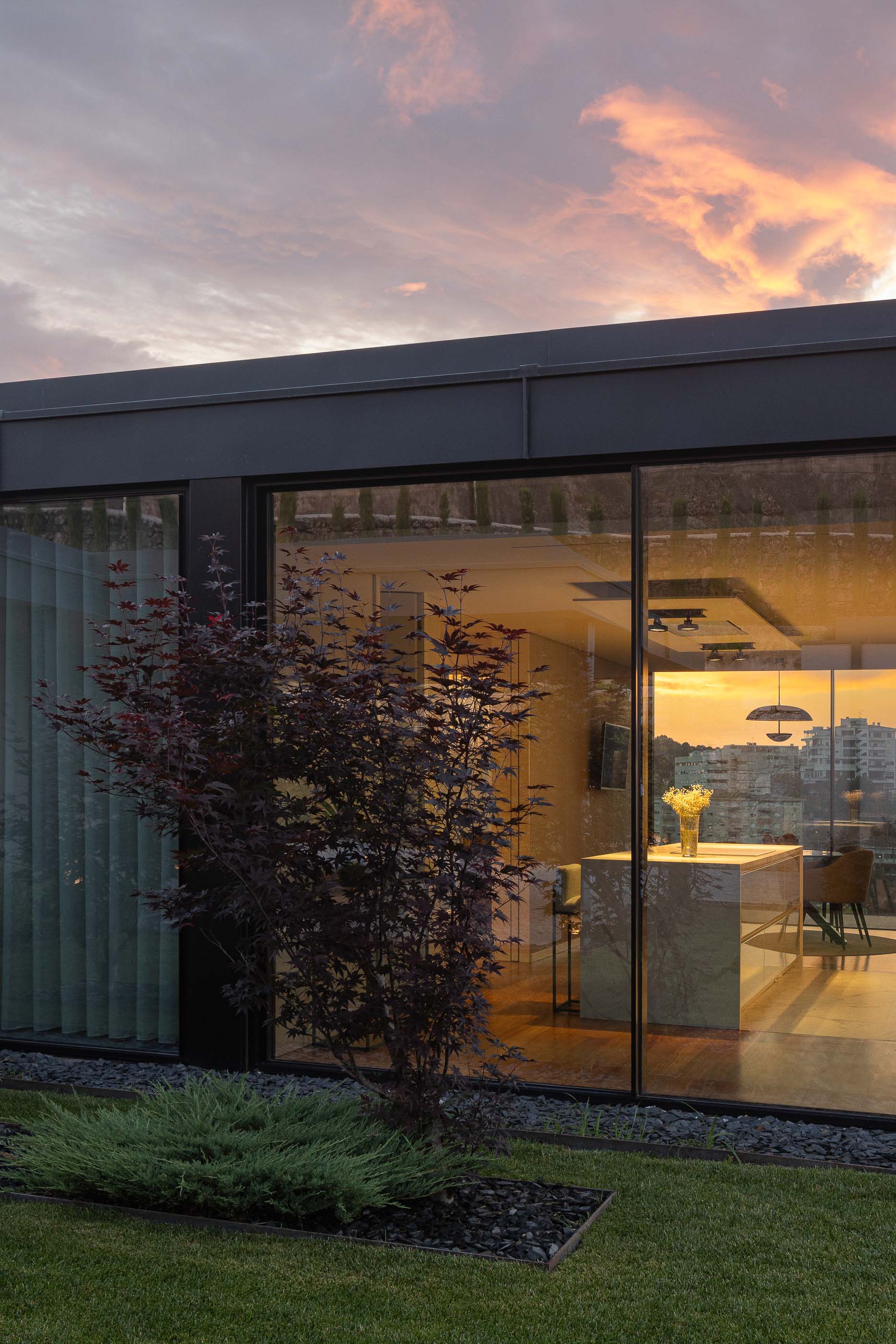 Exterior: Detalhe da janela da cozinha com vista para o jardim e paisagem urbana ao pôr do sol. Arquitetura moderna e integrada na natureza. — Casa da Encosta arquitetura Ricardo Azevedo Arquitecto em Santo Tirso