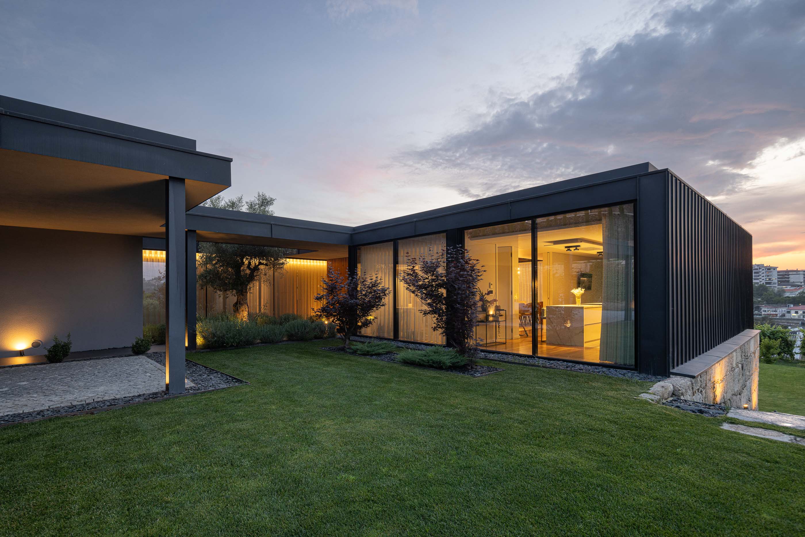 Exterior: Vista da casa com pátio interior, jardim relvado e iluminação noturna. Arquitetura moderna e elegante. — Casa da Encosta arquitetura Ricardo Azevedo Arquitecto em Santo Tirso