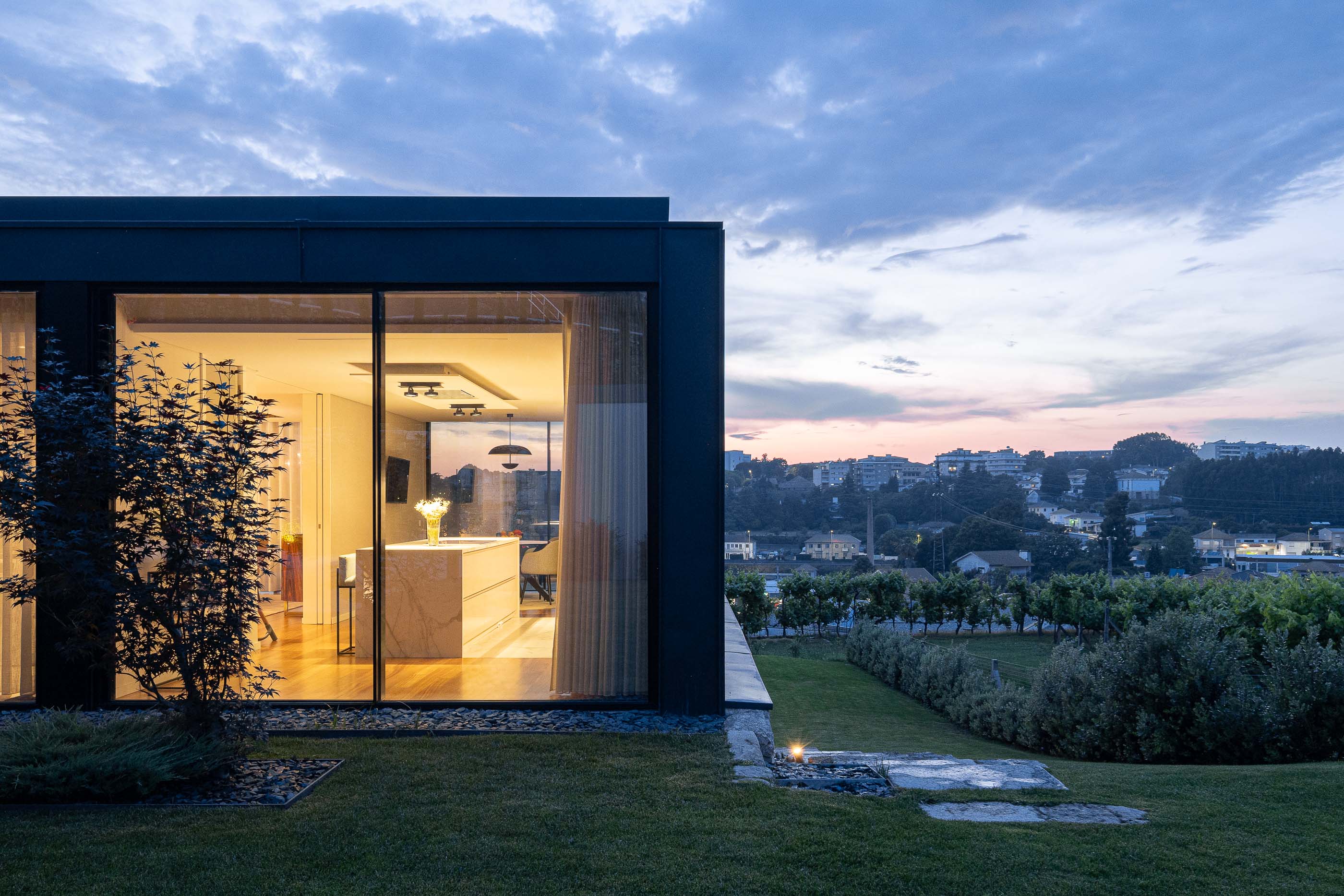 Exterior: Vista da cozinha com iluminação noturna e paisagem urbana ao fundo. Arquitetura moderna e integrada na paisagem. — Casa da Encosta arquitetura Ricardo Azevedo Arquitecto em Santo Tirso