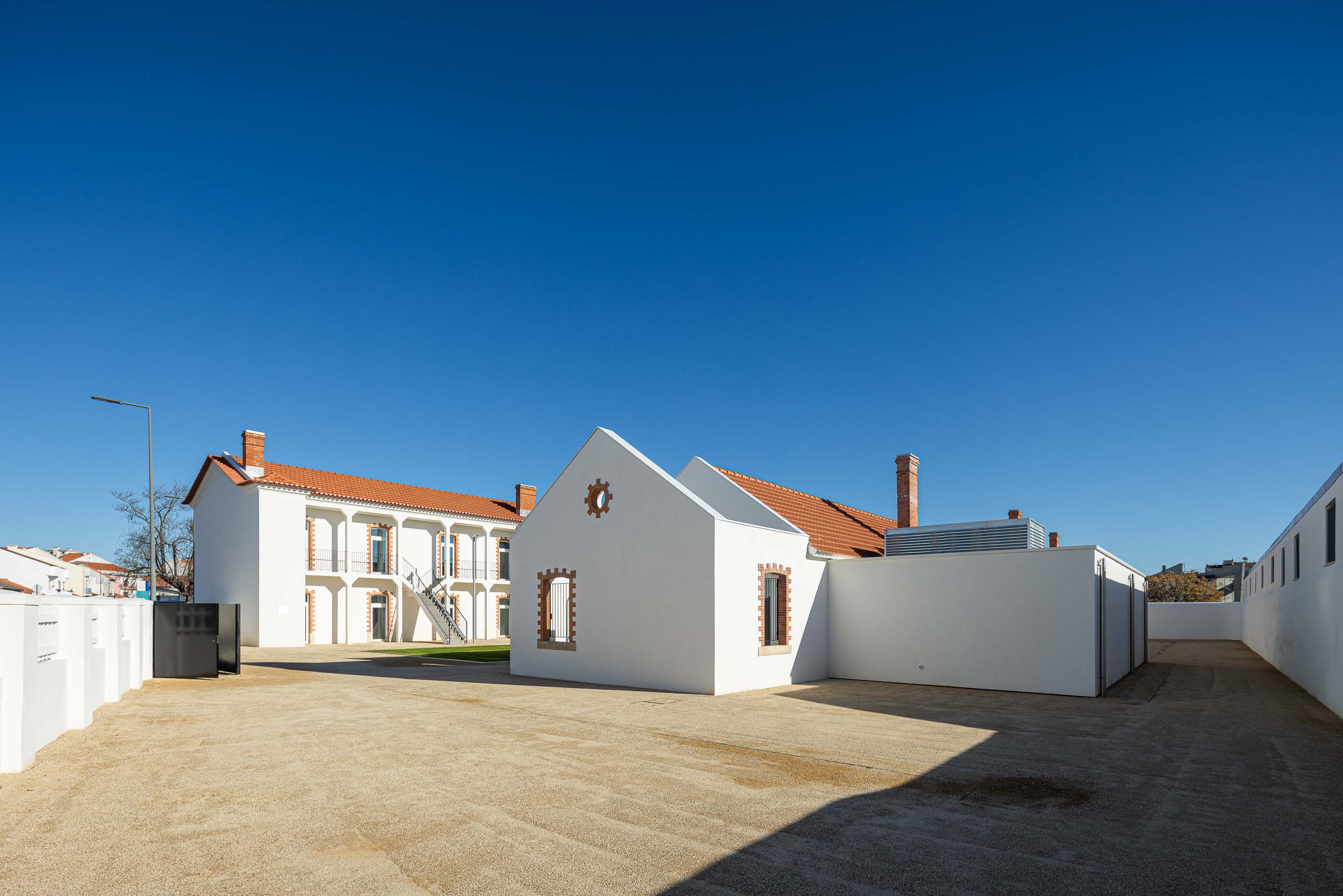 Exterior: Pátio com gravilha, edifícios brancos com telhado cerâmico e detalhes em tijolo aparente. — Reabilitação Urbana do Bairro do Boneco arquitetura Adarq - André David Arquitecto em Entroncamento