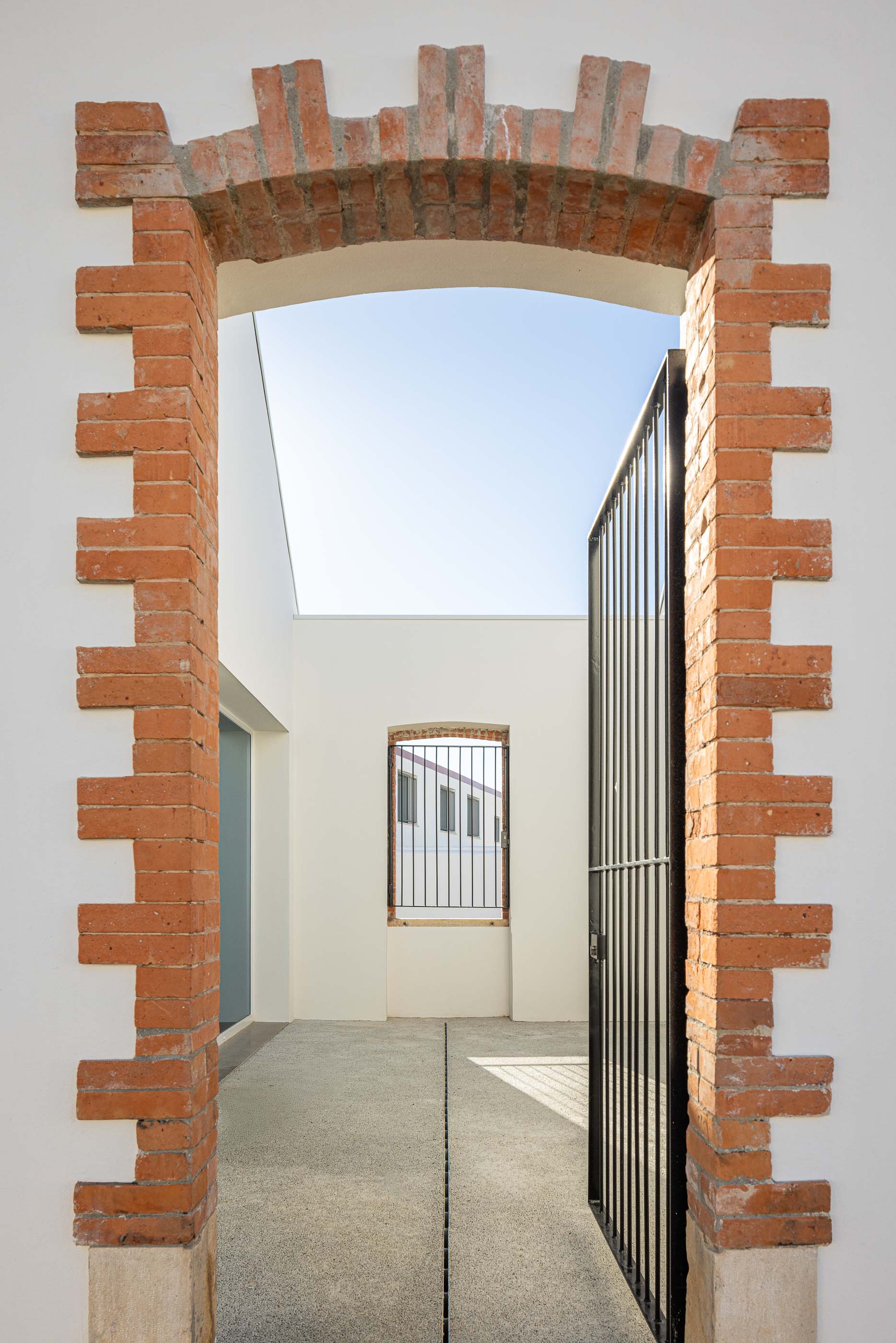 Exterior: Arco em tijolo aparente, com portão metálico, pavimento em betão e vista para o pátio interior. — Reabilitação Urbana do Bairro do Boneco arquitetura Adarq - André David Arquitecto em Entroncamento