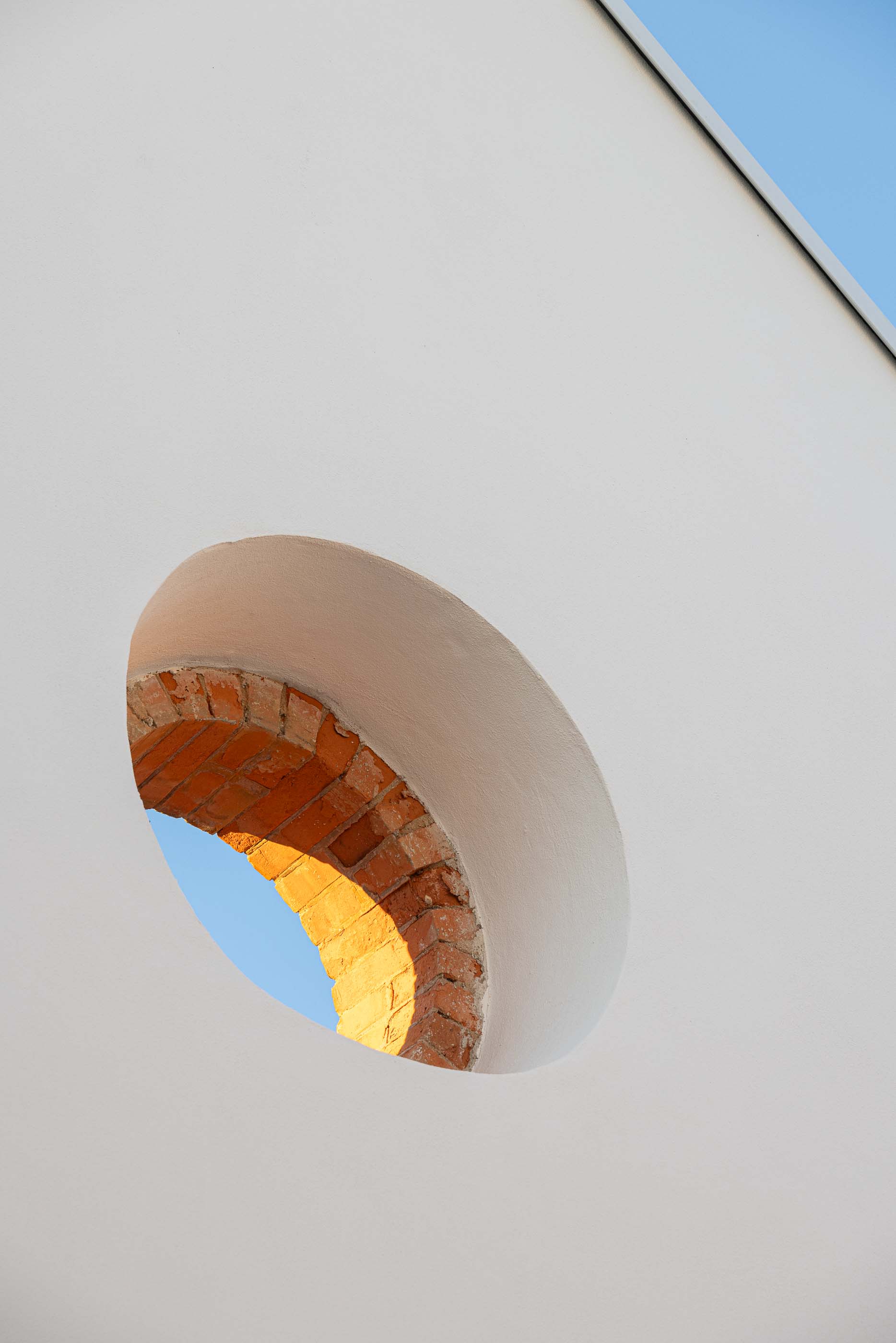 Detalhe: Óculo circular em tijolo aparente numa fachada branca, com céu azul no fundo. — Reabilitação Urbana do Bairro do Boneco arquitetura Adarq - André David Arquitecto em Entroncamento