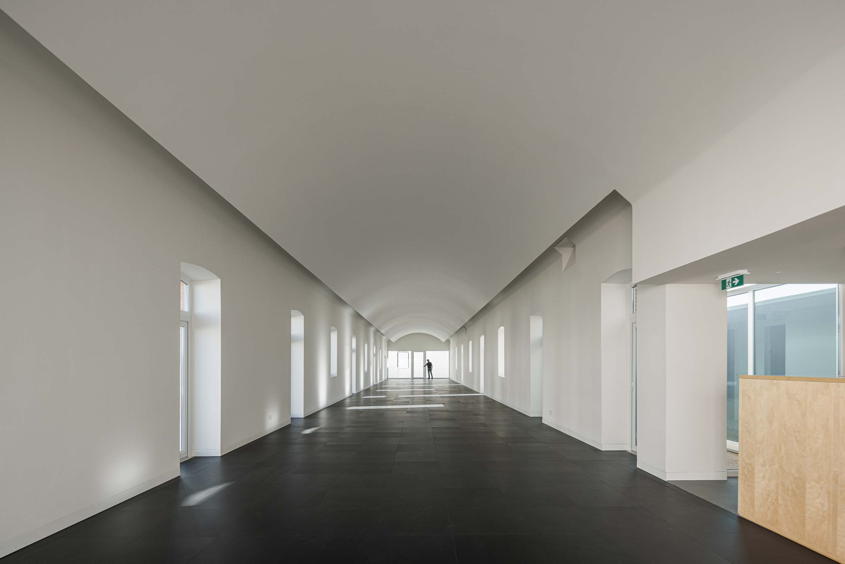 Espaço amplo: Salão comprido com pavimento escuro, paredes brancas e janelas ao longo do corredor. — Reabilitação Urbana do Bairro do Boneco arquitetura Adarq - André David Arquitecto em Entroncamento