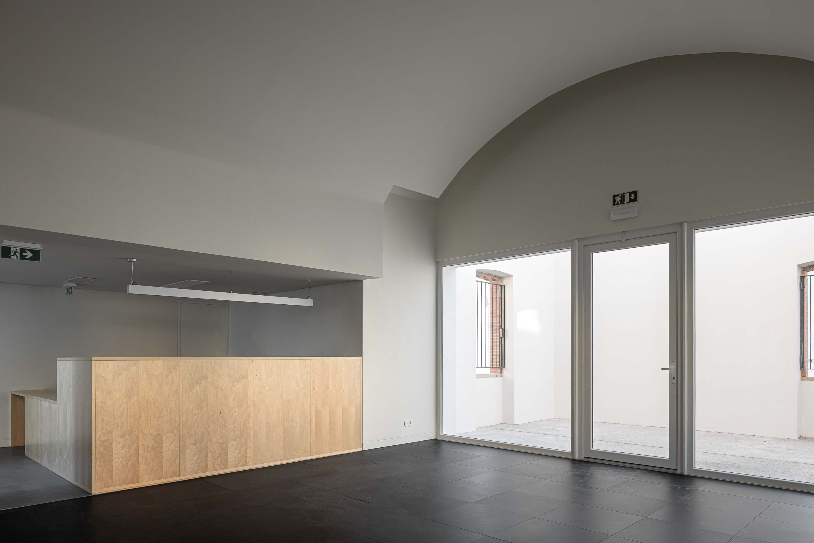 Espaço amplo: Zona de receção com balcão em madeira clara, pavimento escuro e acesso ao pátio exterior. — Reabilitação Urbana do Bairro do Boneco arquitetura Adarq - André David Arquitecto em Entroncamento