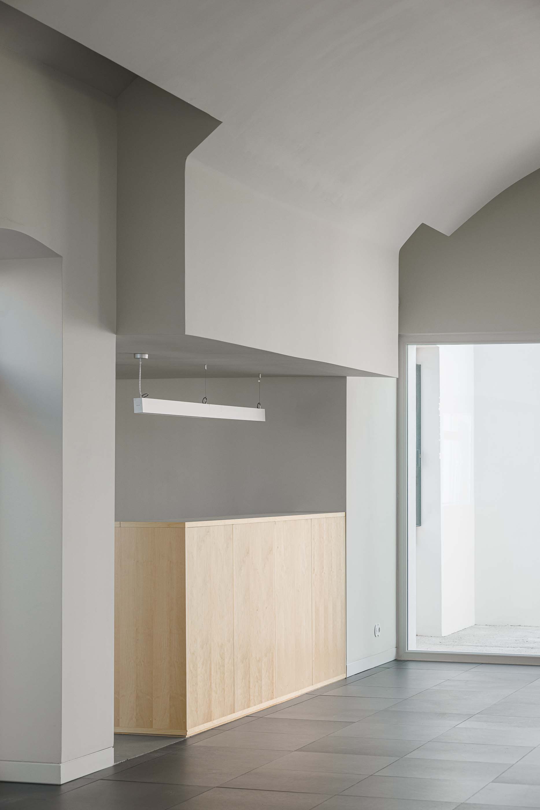 Espaço amplo: Balcão em madeira clara, pavimento escuro e iluminação suspensa linear. — Reabilitação Urbana do Bairro do Boneco arquitetura Adarq - André David Arquitecto em Entroncamento