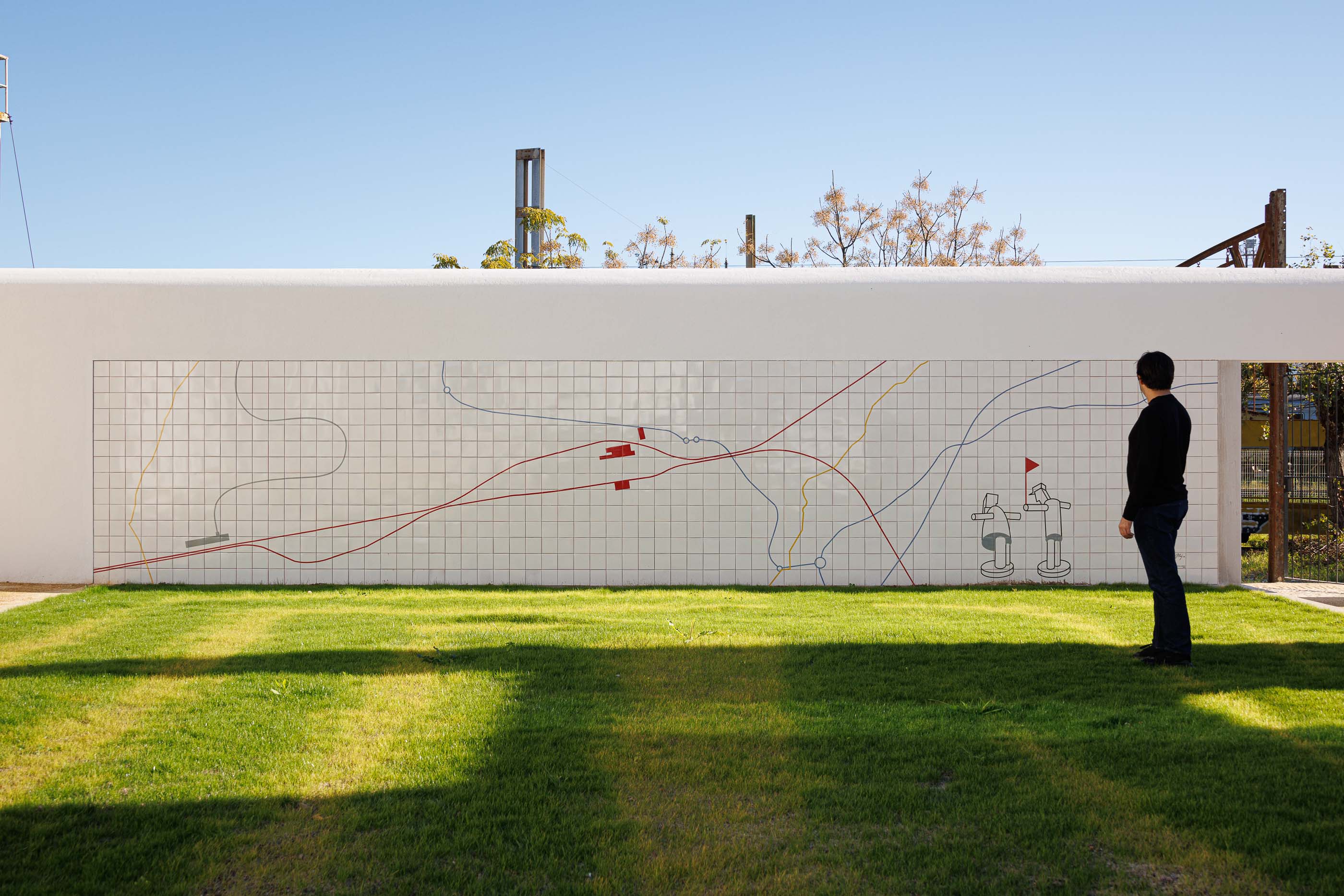 Exterior: Muro revestido com azulejos brancos e desenho linear a cores, com relvado e pessoa a observar. — Reabilitação Urbana do Bairro do Boneco arquitetura Adarq - André David Arquitecto em Entroncamento