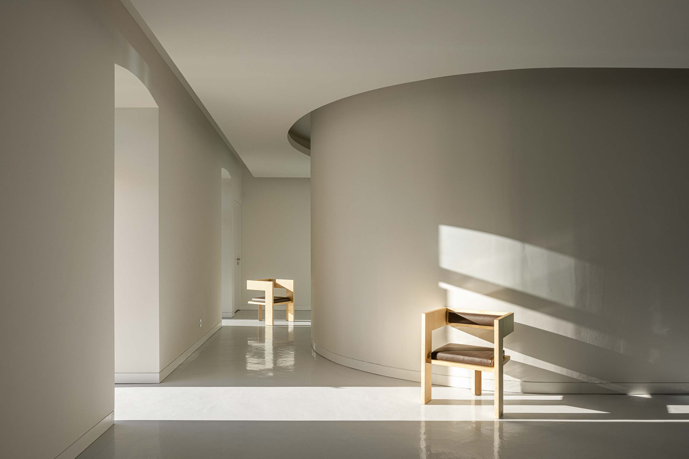 Corredor: Espaço interior com paredes curvas, pavimento claro, cadeiras de madeira e iluminação natural. — Reabilitação Urbana do Bairro do Boneco arquitetura Adarq - André David Arquitecto em Entroncamento