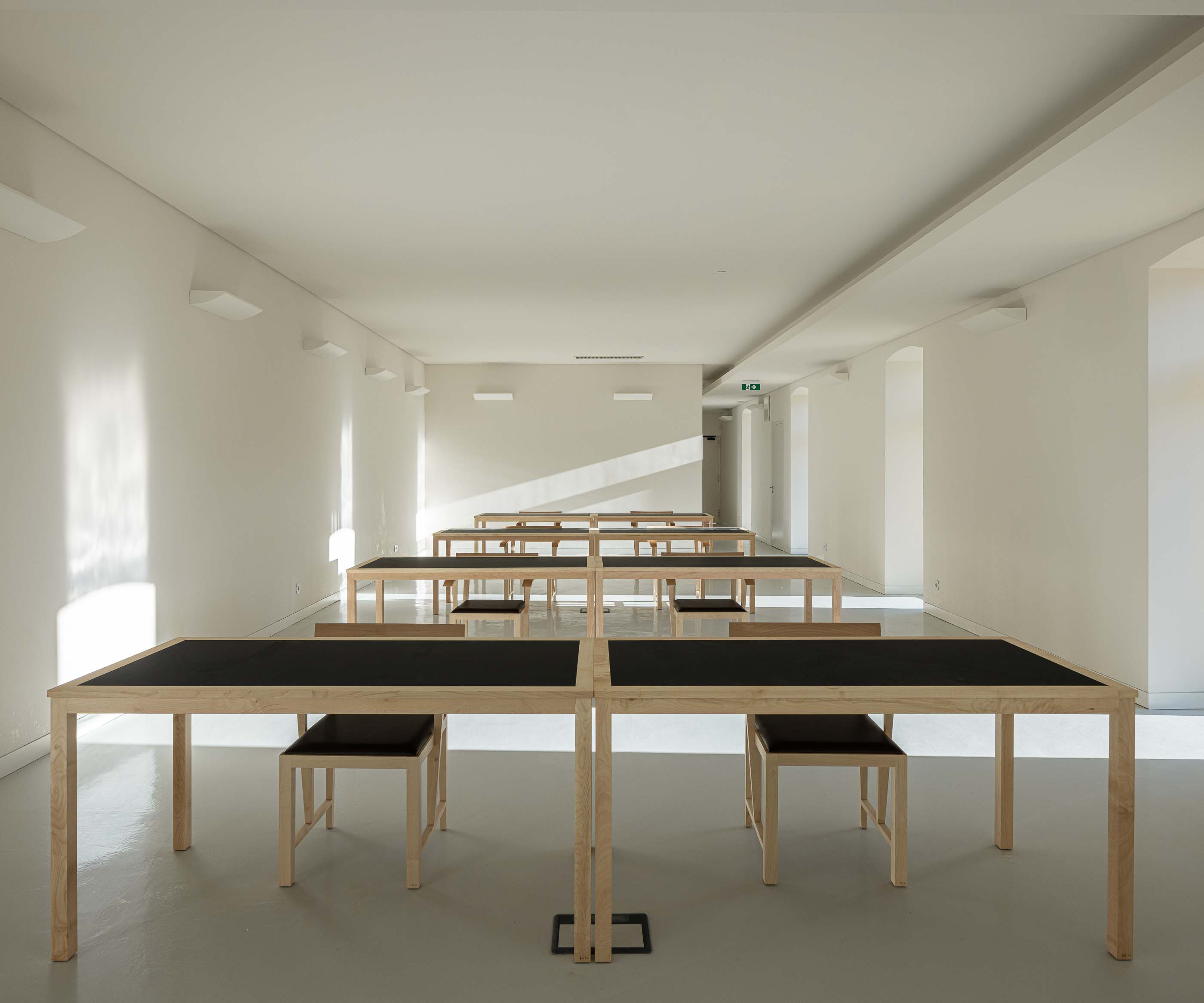 Sala: Espaço amplo com mesas de trabalho em madeira e cadeiras, pavimento claro e iluminação linear. — Reabilitação Urbana do Bairro do Boneco arquitetura Adarq - André David Arquitecto em Entroncamento
