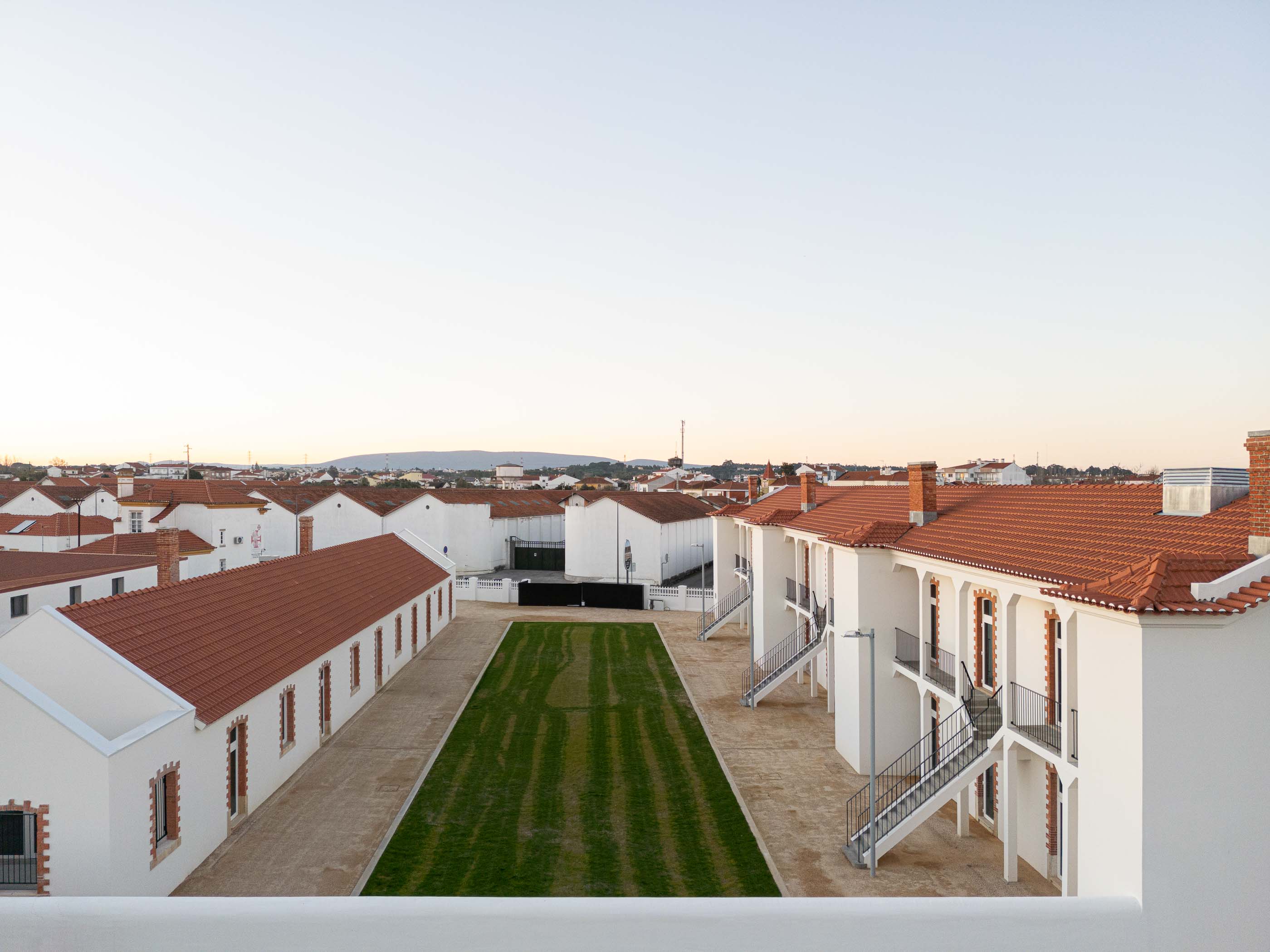 Vista aérea: Conjunto habitacional branco com telhados vermelhos e pátio relvado central. — Reabilitação Urbana do Bairro do Boneco arquitetura Adarq - André David Arquitecto em Entroncamento