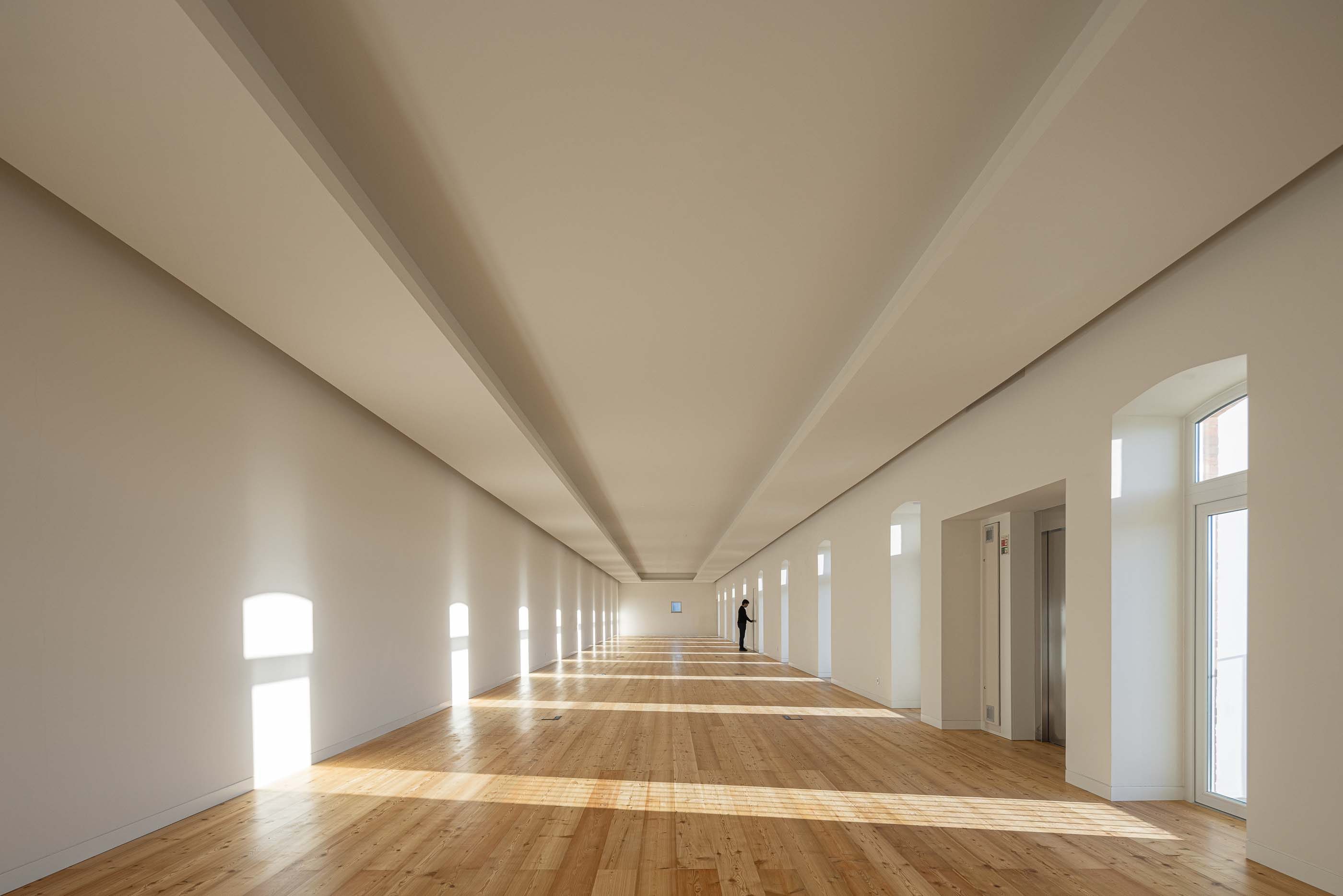 Espaço amplo: Salão comprido com pavimento em madeira, paredes brancas e janelas ao longo do corredor. — Reabilitação Urbana do Bairro do Boneco arquitetura Adarq - André David Arquitecto em Entroncamento