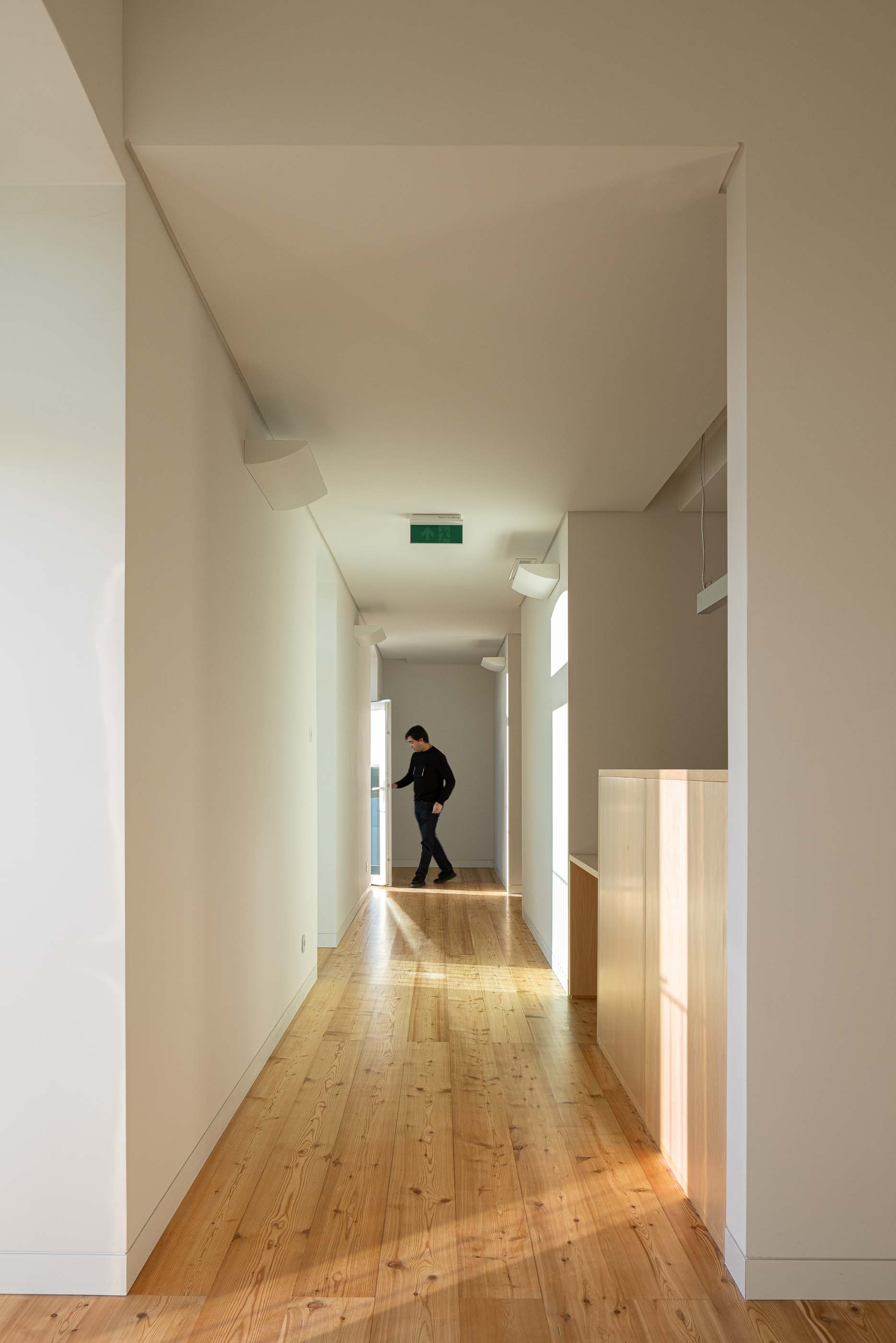 Corredor: Espaço interior com pavimento em madeira, paredes brancas e iluminação linear suspensa. — Reabilitação Urbana do Bairro do Boneco arquitetura Adarq - André David Arquitecto em Entroncamento