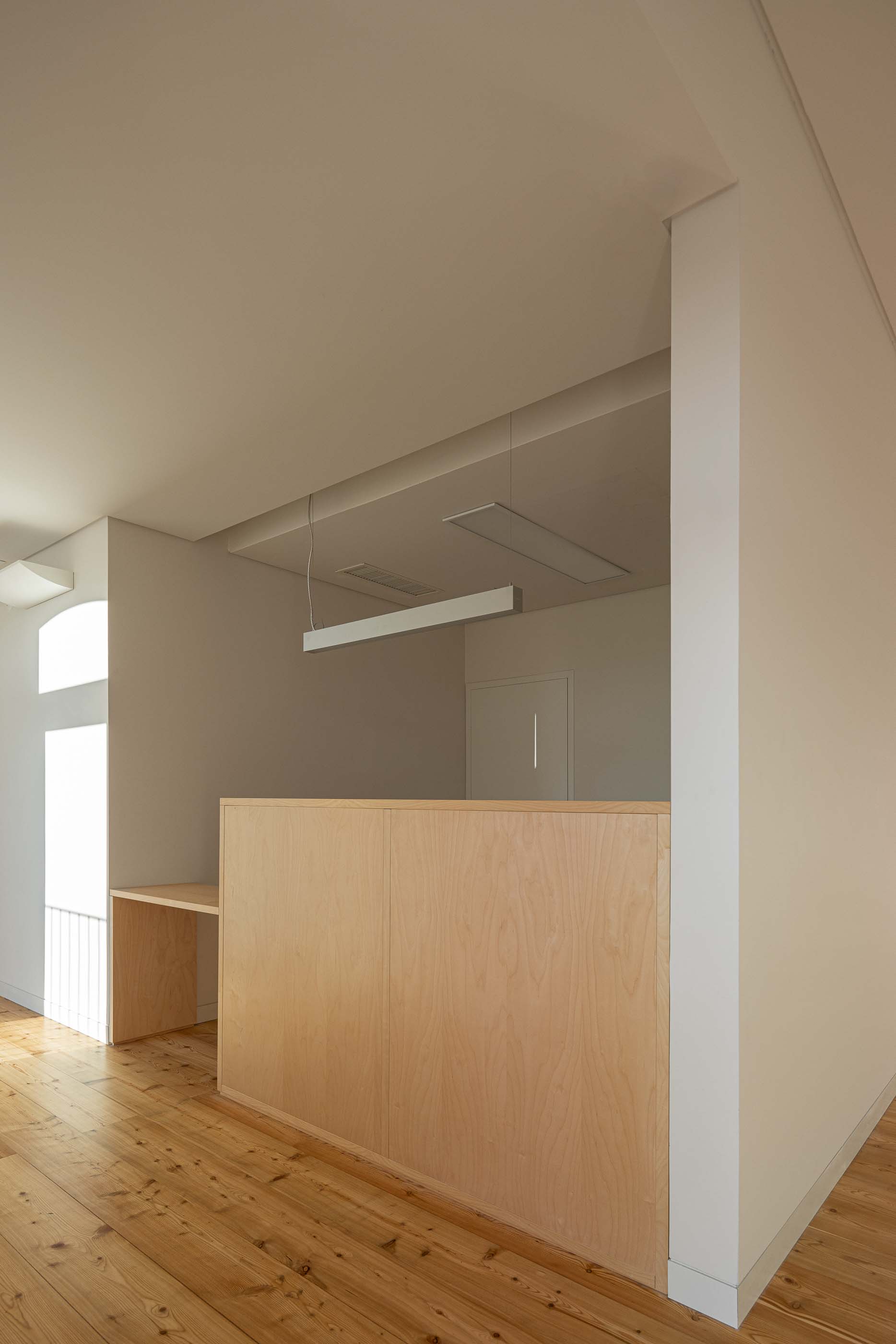 Espaço amplo: Balcão em madeira clara, pavimento em madeira e iluminação linear suspensa. — Reabilitação Urbana do Bairro do Boneco arquitetura Adarq - André David Arquitecto em Entroncamento