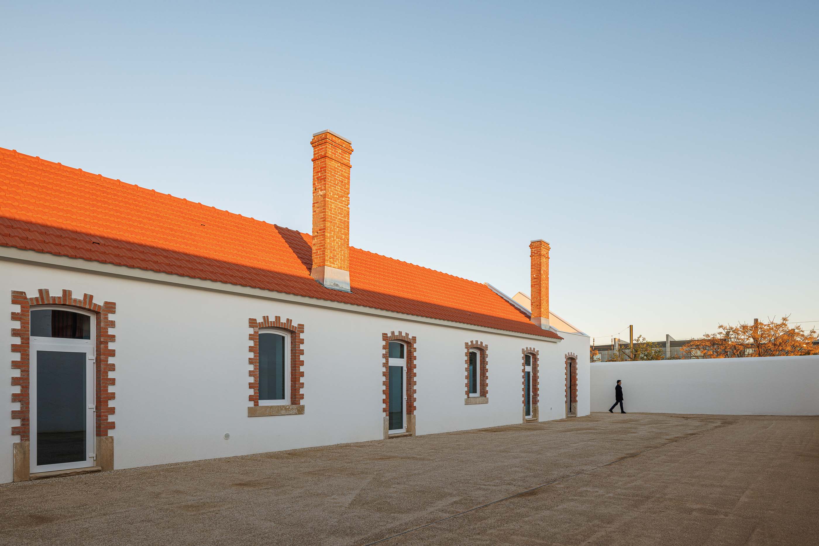 Exterior: Edifício branco com telhado cerâmico e chaminés em tijolo, pátio com gravilha. — Reabilitação Urbana do Bairro do Boneco arquitetura Adarq - André David Arquitecto em Entroncamento