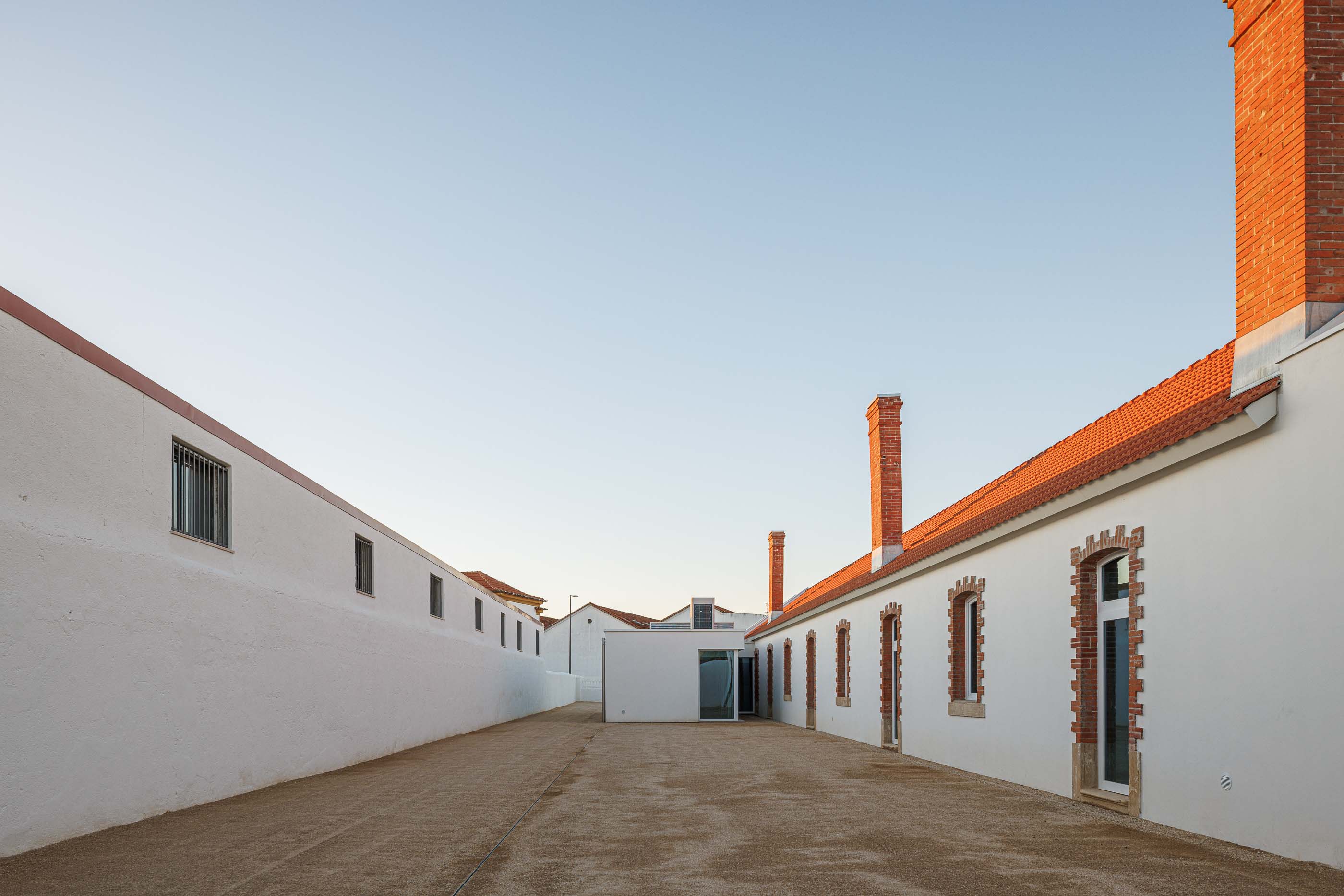 Exterior: Pátio com gravilha, edifícios brancos com telhado cerâmico e chaminés em tijolo. — Reabilitação Urbana do Bairro do Boneco arquitetura Adarq - André David Arquitecto em Entroncamento