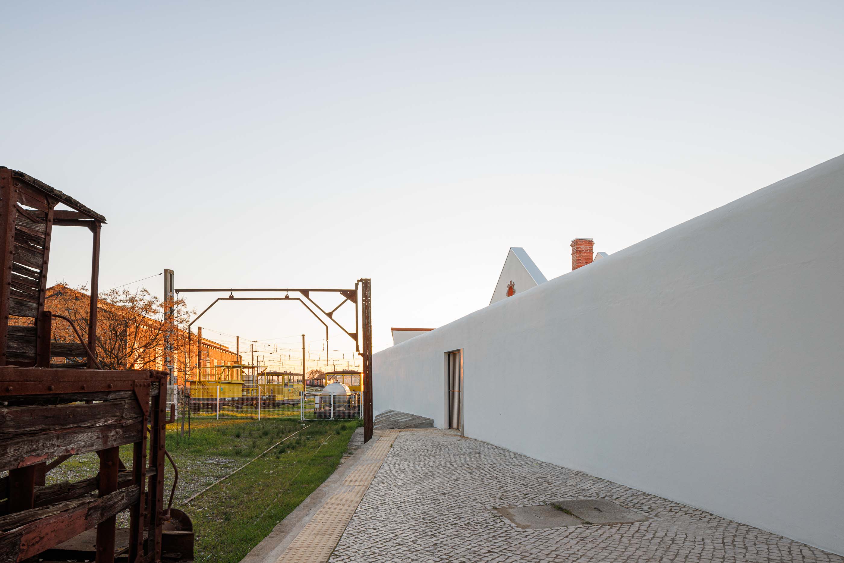 Exterior: Muro branco, elementos ferroviários antigos e edifícios brancos com telhado cerâmico. — Reabilitação Urbana do Bairro do Boneco arquitetura Adarq - André David Arquitecto em Entroncamento