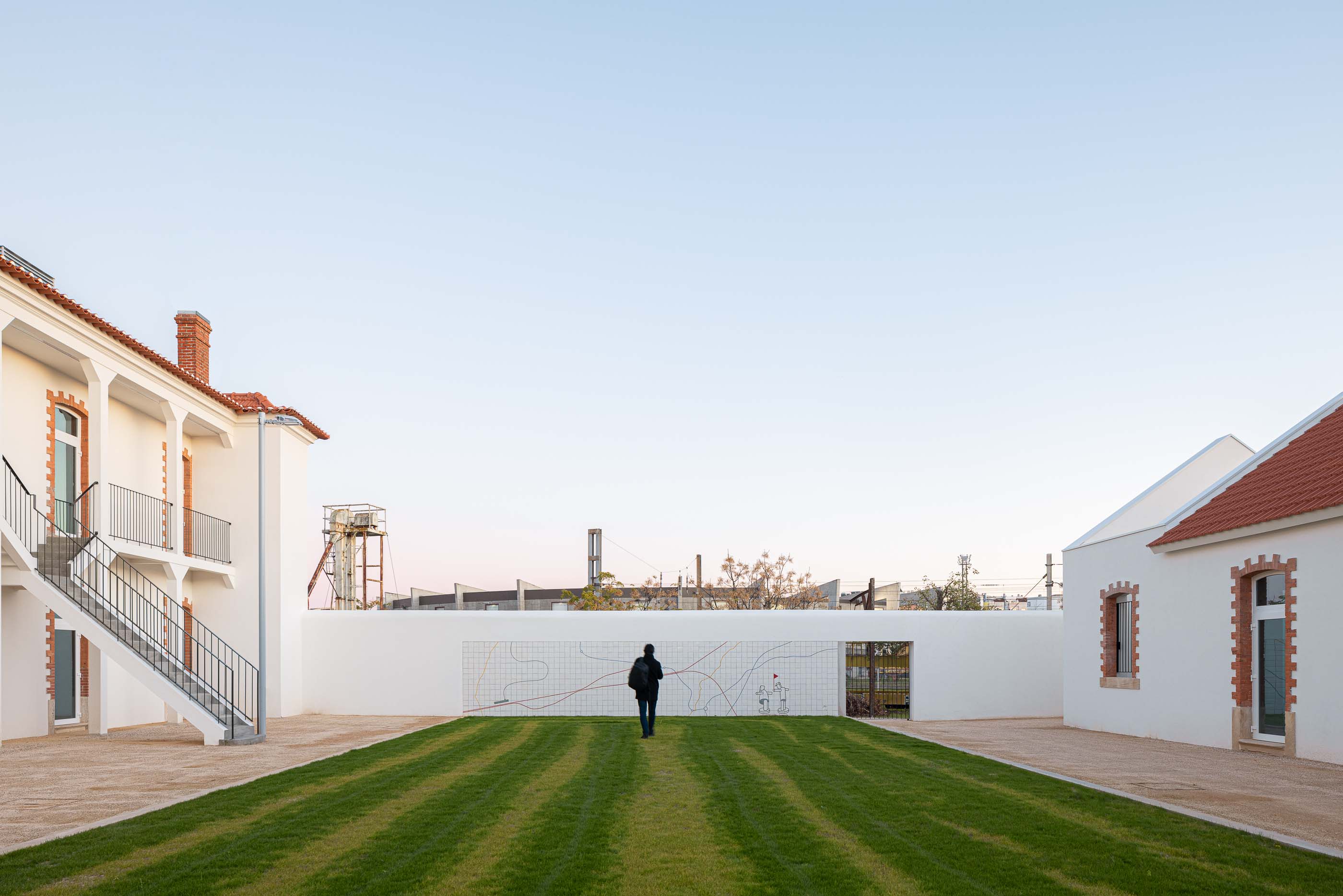 Exterior: Pátio relvado, edifícios brancos com telhado cerâmico e muro com revestimento cerâmico. — Reabilitação Urbana do Bairro do Boneco arquitetura Adarq - André David Arquitecto em Entroncamento