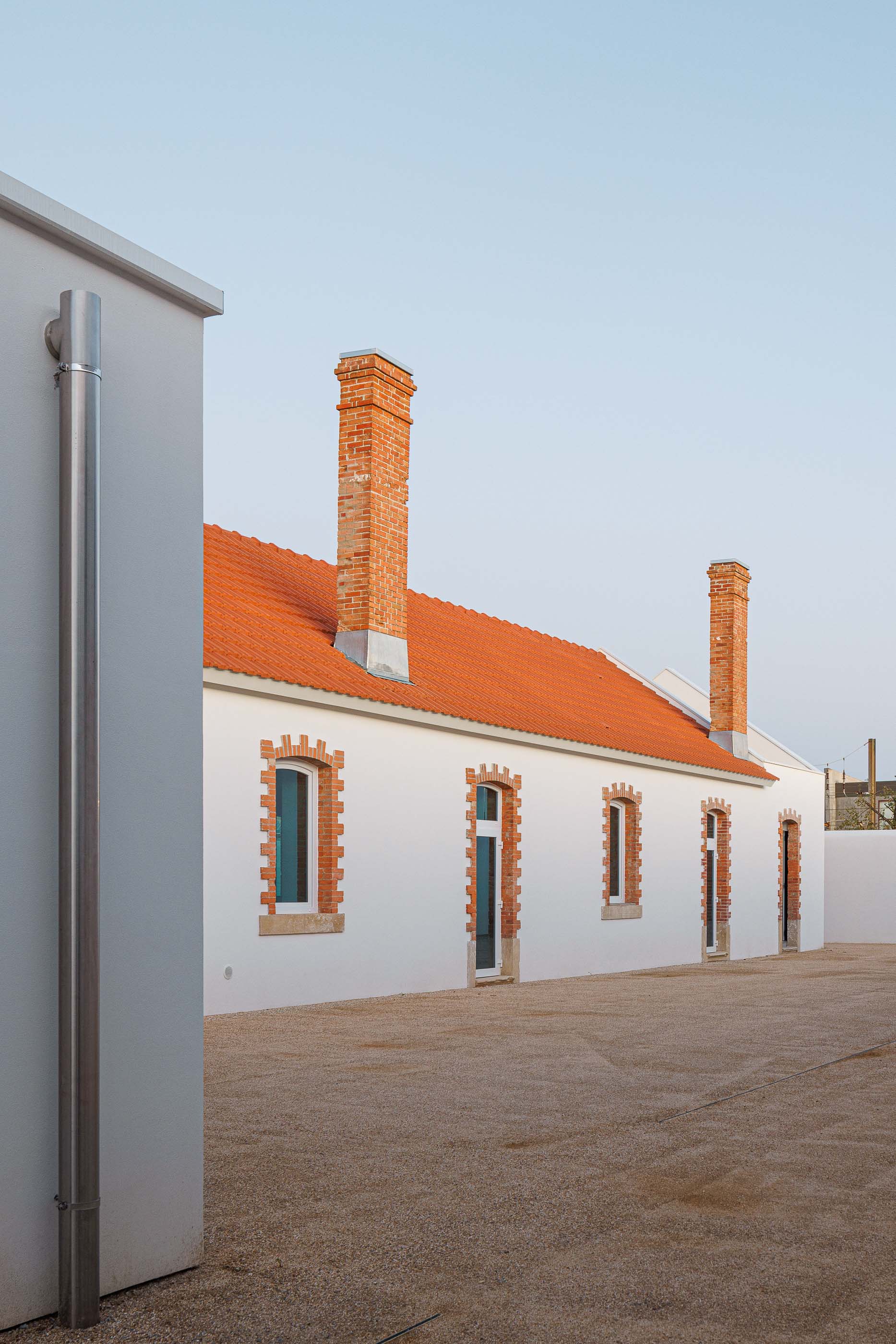 Exterior: Edifício branco com telhado cerâmico e chaminés em tijolo, pátio com gravilha. — Reabilitação Urbana do Bairro do Boneco arquitetura Adarq - André David Arquitecto em Entroncamento
