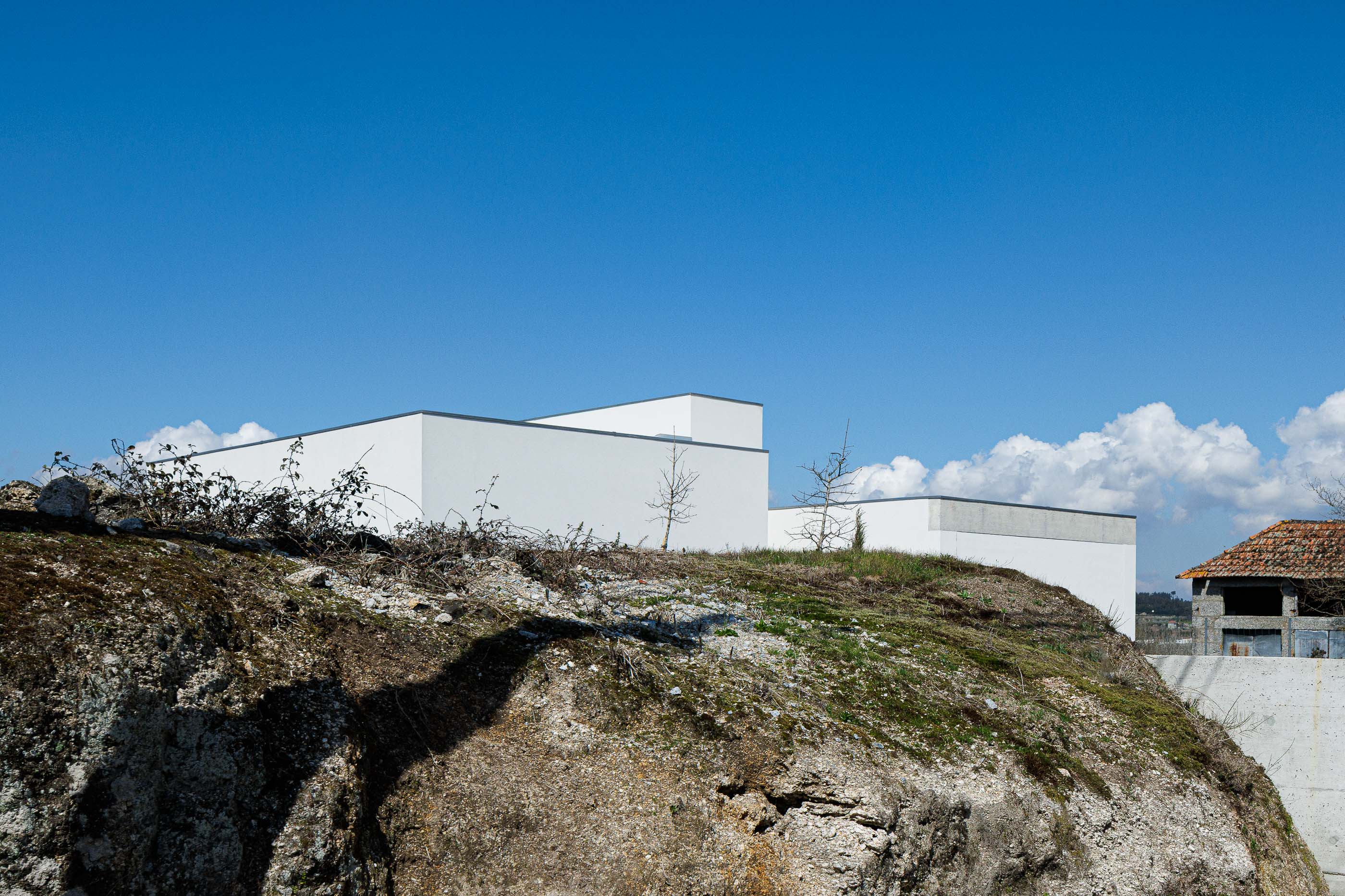 Exterior: Vista da casa branca de arquitetura minimalista, implantada num terreno rochoso e natural. — Pedrantil House arquitetura Helder da Rocha Arquitectos em Penafiel